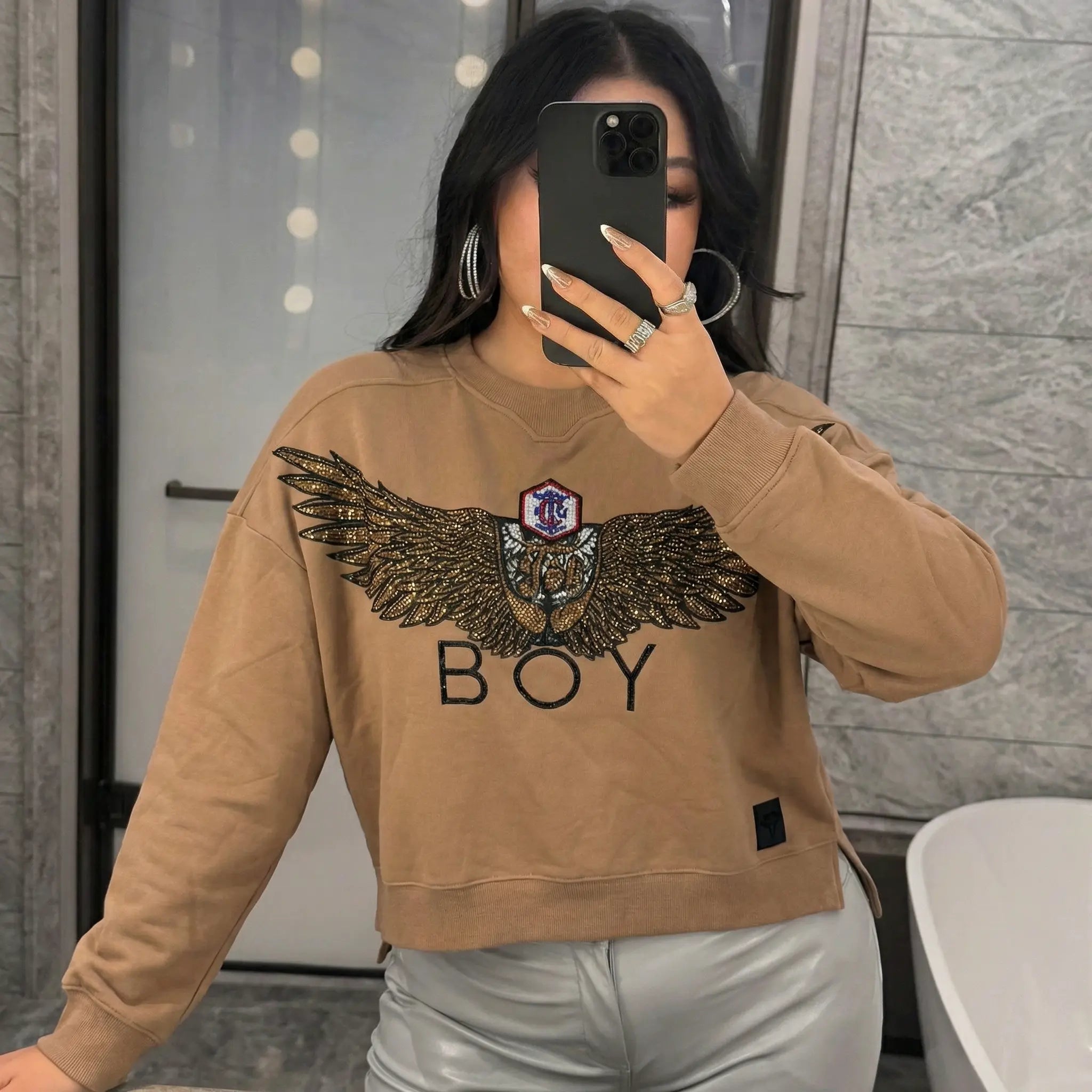Person wearing a tan sweatshirt with a logo, taking a mirror selfie. Rhinestone Wing Logo Sweatshirt