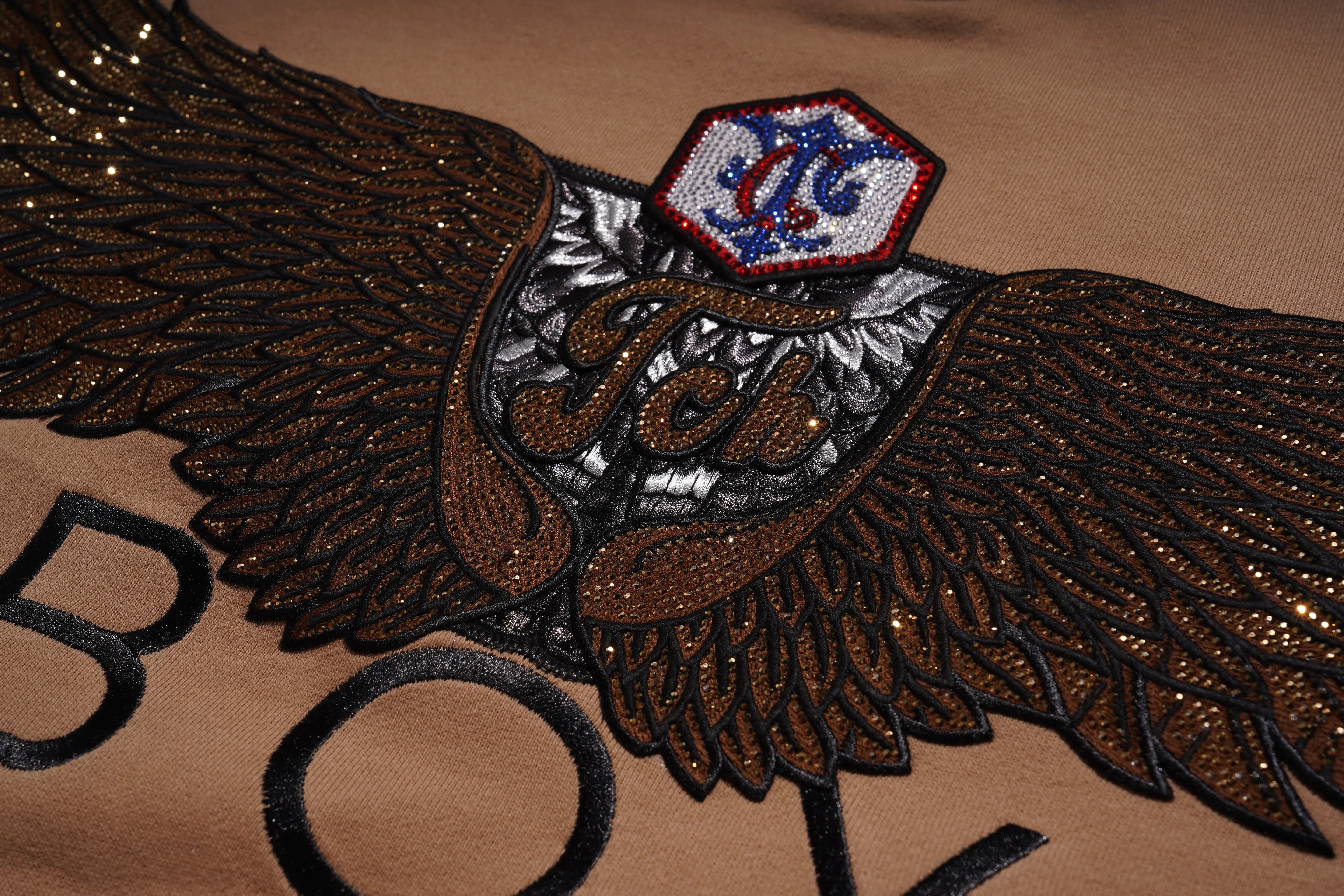 Close-up of a decorative patch with wings and a logo on a brown background， Rhinestone Wing Logo Sweatshirt