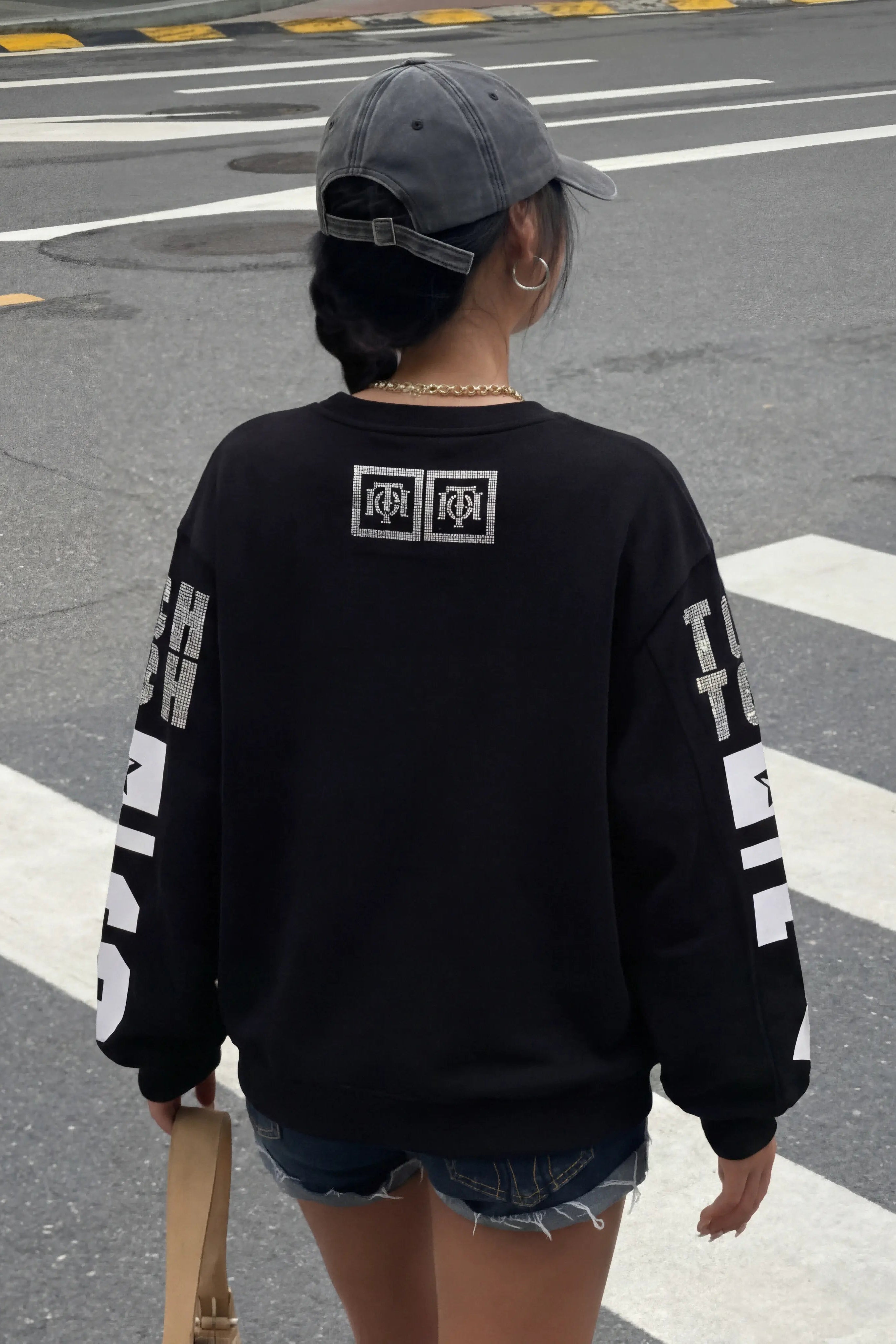 Person wearing a black sweatshirt with white text and logos, standing on a crosswalk. TCH 1976 Rhinestone Wings Sweatshirt