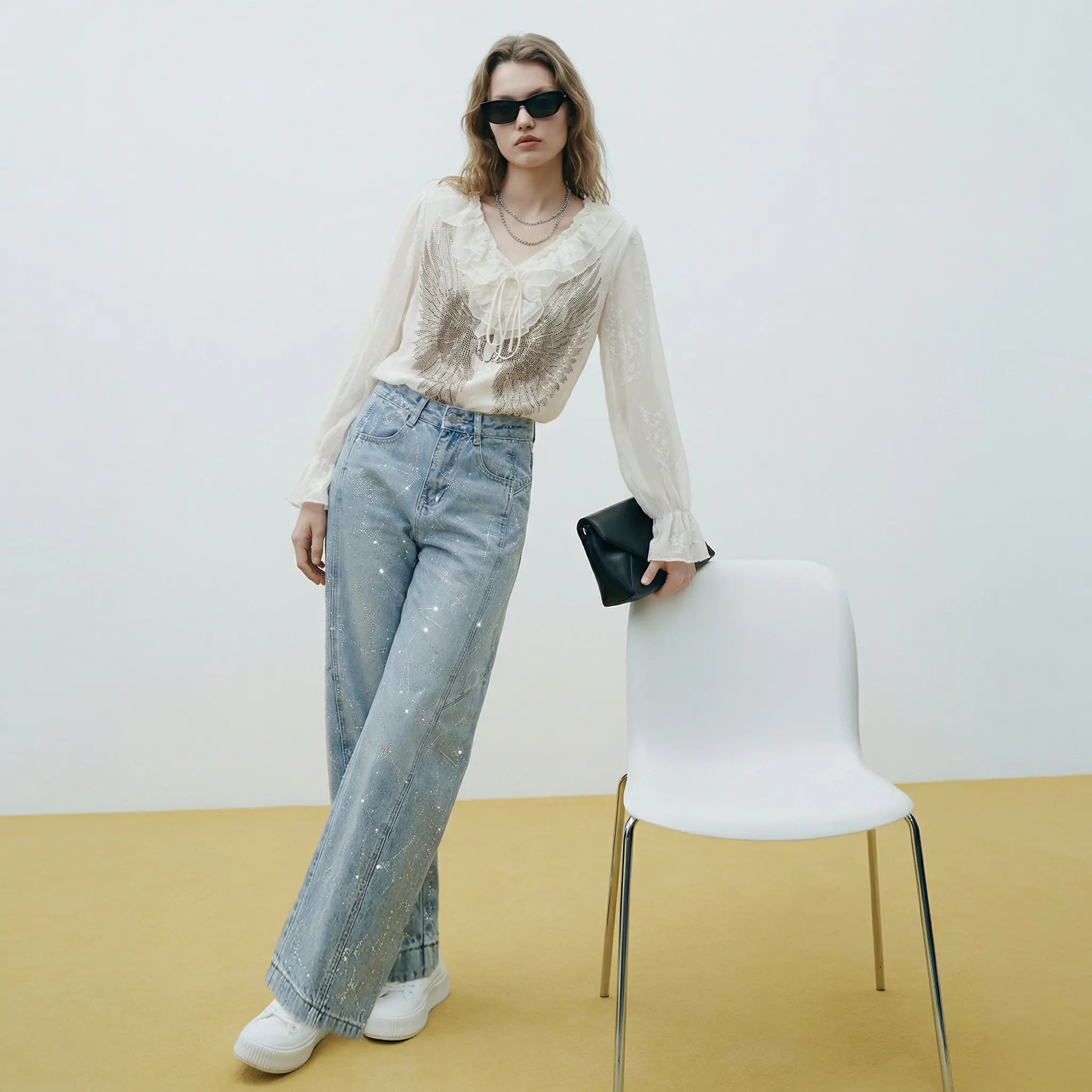 Woman in a stylish outfit with jeans and a top, standing next to a white chair against a plain background. TCH Angel Wings Rhinestone Ruffle Blouse