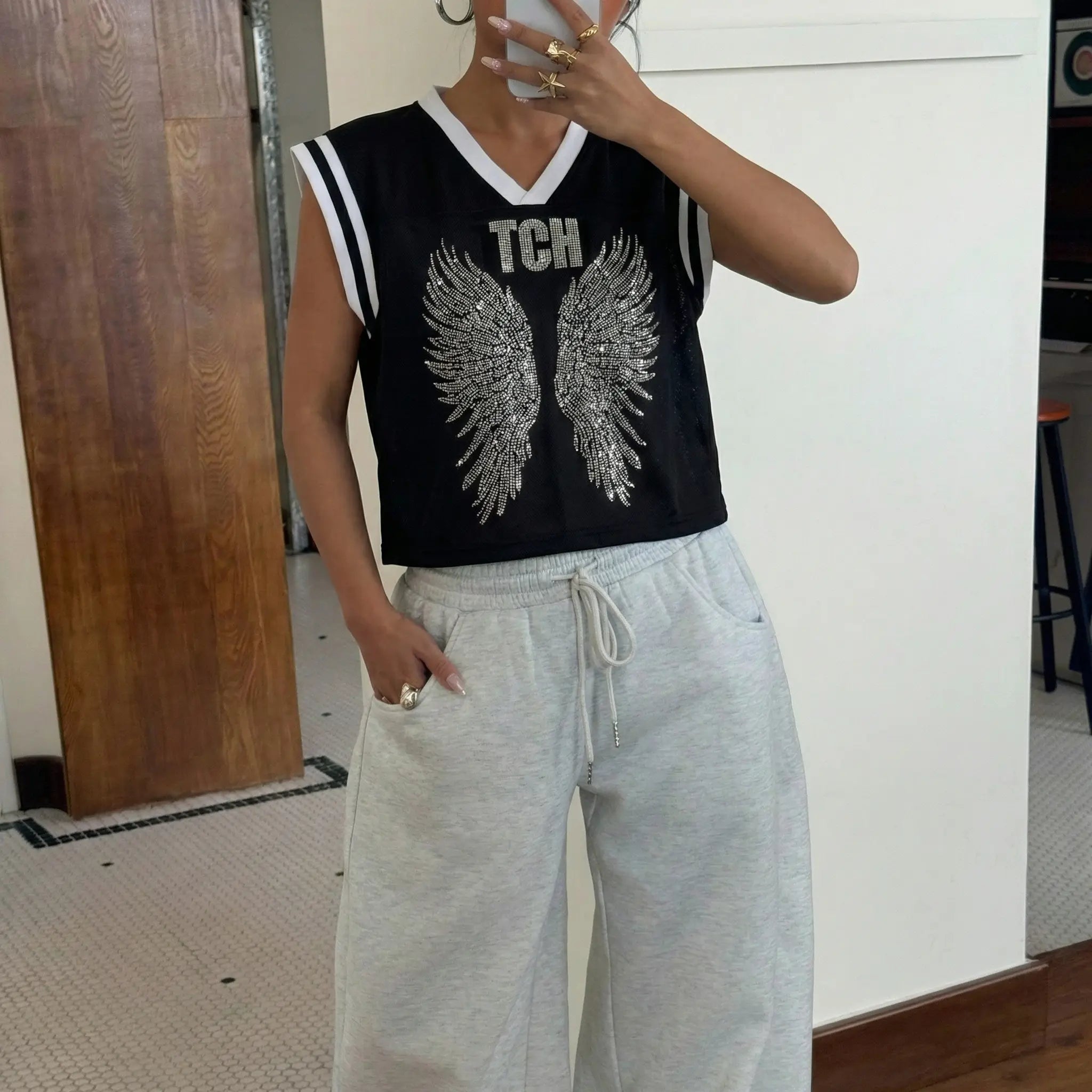 Person wearing a black sleeveless top with silver wings and 'TCH' letters, paired with light gray sweatpants. TCH Angel Wings Rhinestone V-Neck Tee