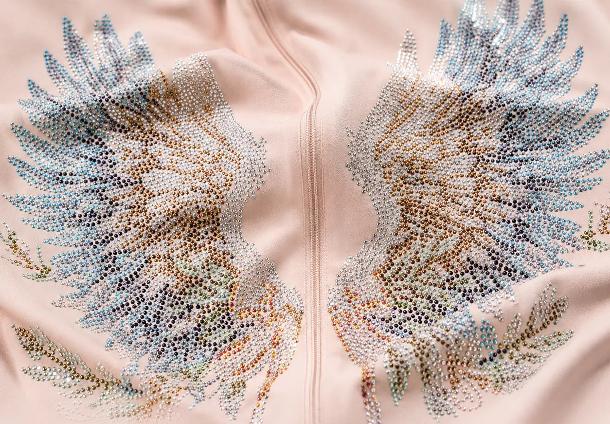 Close-up of a light blush pink garment featuring large symmetrical wing motifs covered in iridescent rhinestones that shift from pale blue to champagne, gold, and rose-gold tones. TCH Angel Wings Rhinestone Zip-Up Hoodie