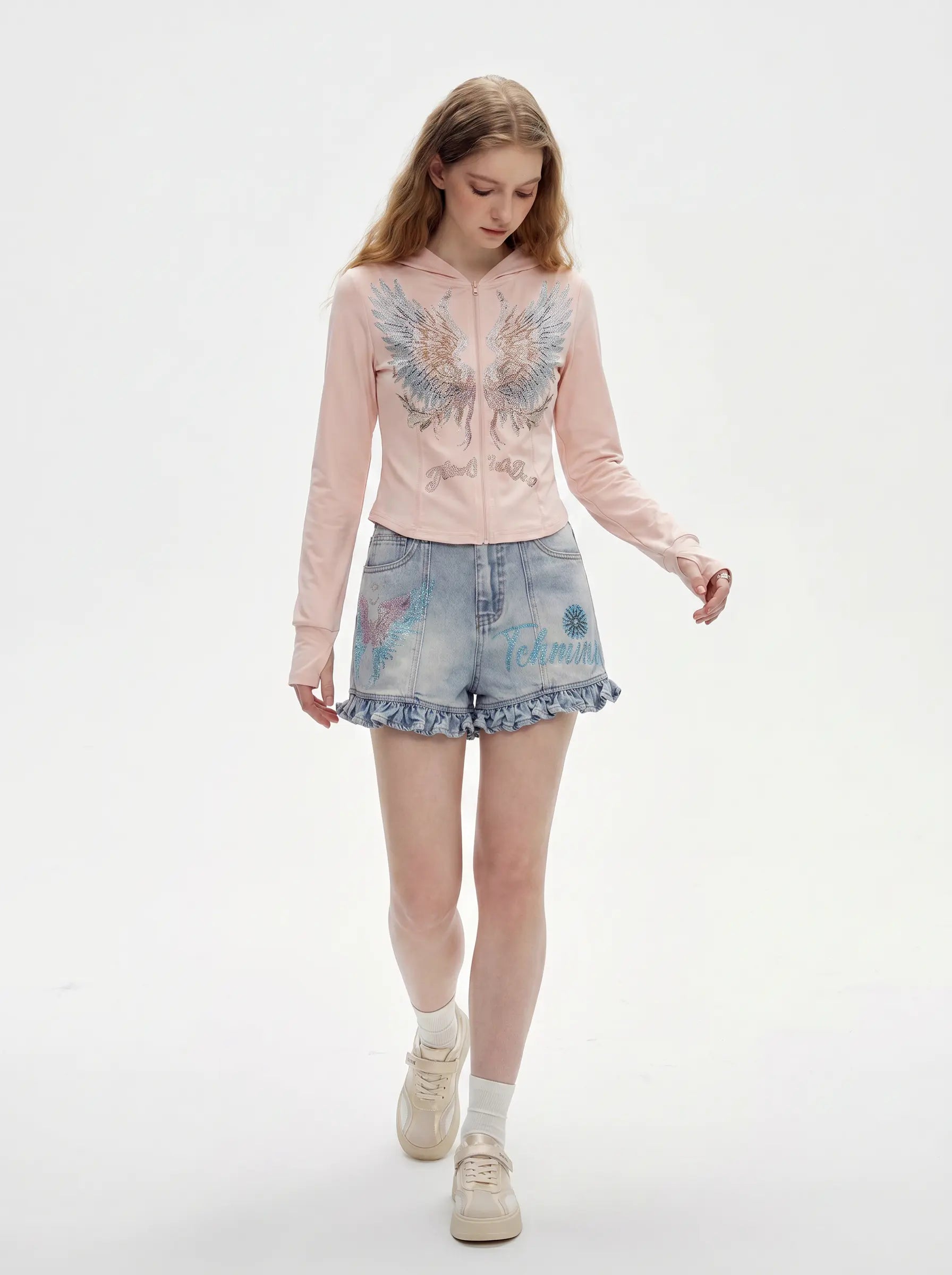 A young woman walking against a plain white studio background, wearing a light pink zip-up hoodie with a large embroidered angel wings design on the front, light-wash denim shorts, white socks, and beige sneakers. TCH Angel Wings Rhinestone Zip-Up Hoodie