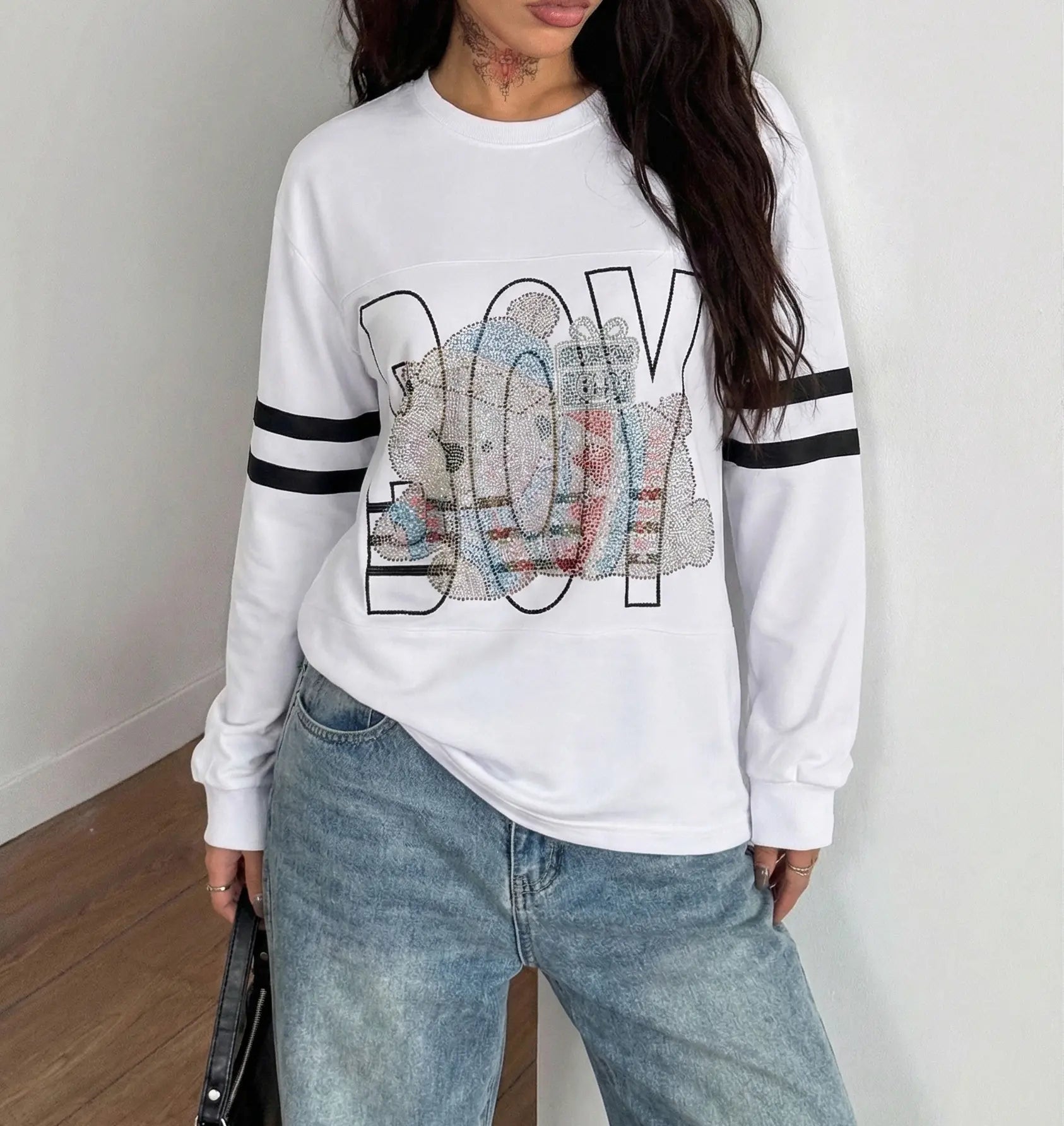 Person wearing a white sweatshirt with colorful 'BOY' design and blue jeans indoors. TCH BOY Rhinestone Bear Sweatshirt-