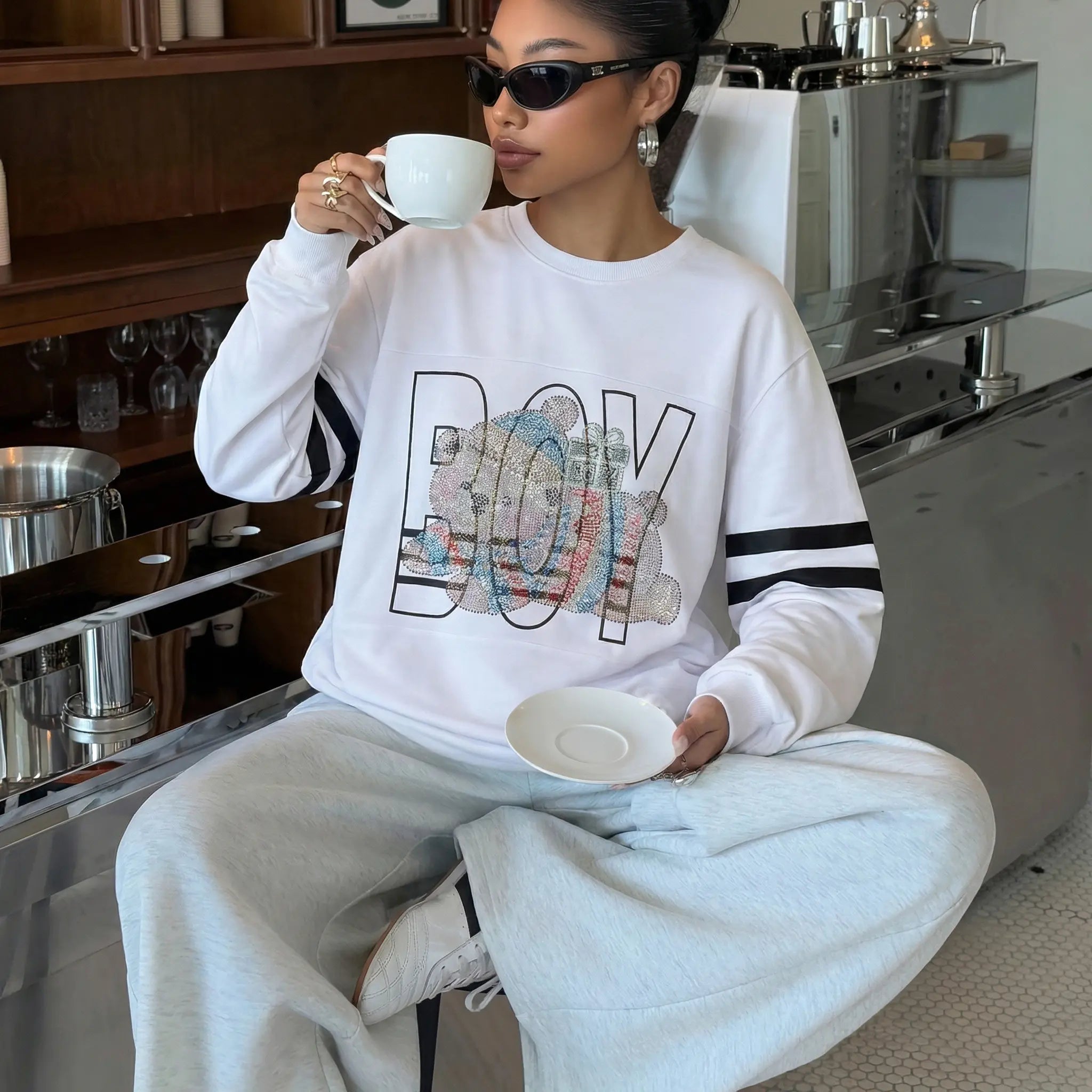 Person wearing a white sweatshirt with colorful text and holding a cup in a kitchen setting. TCH BOY Rhinestone Bear Sweatshirt-