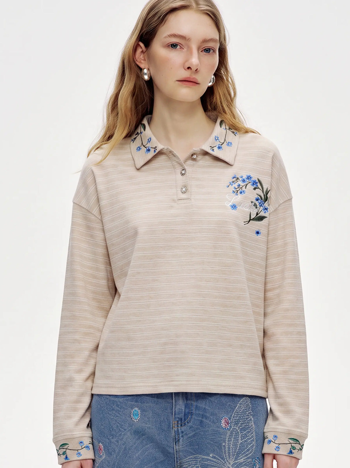 A model wearing a light blue denim jacket with various embroidered patches, including a teddy bear on the left chest and cherries on the sleeve, styled over a white t-shirt and dark pants. TCH Beige Floral Embroidered Long Sleeve Polo Shirt