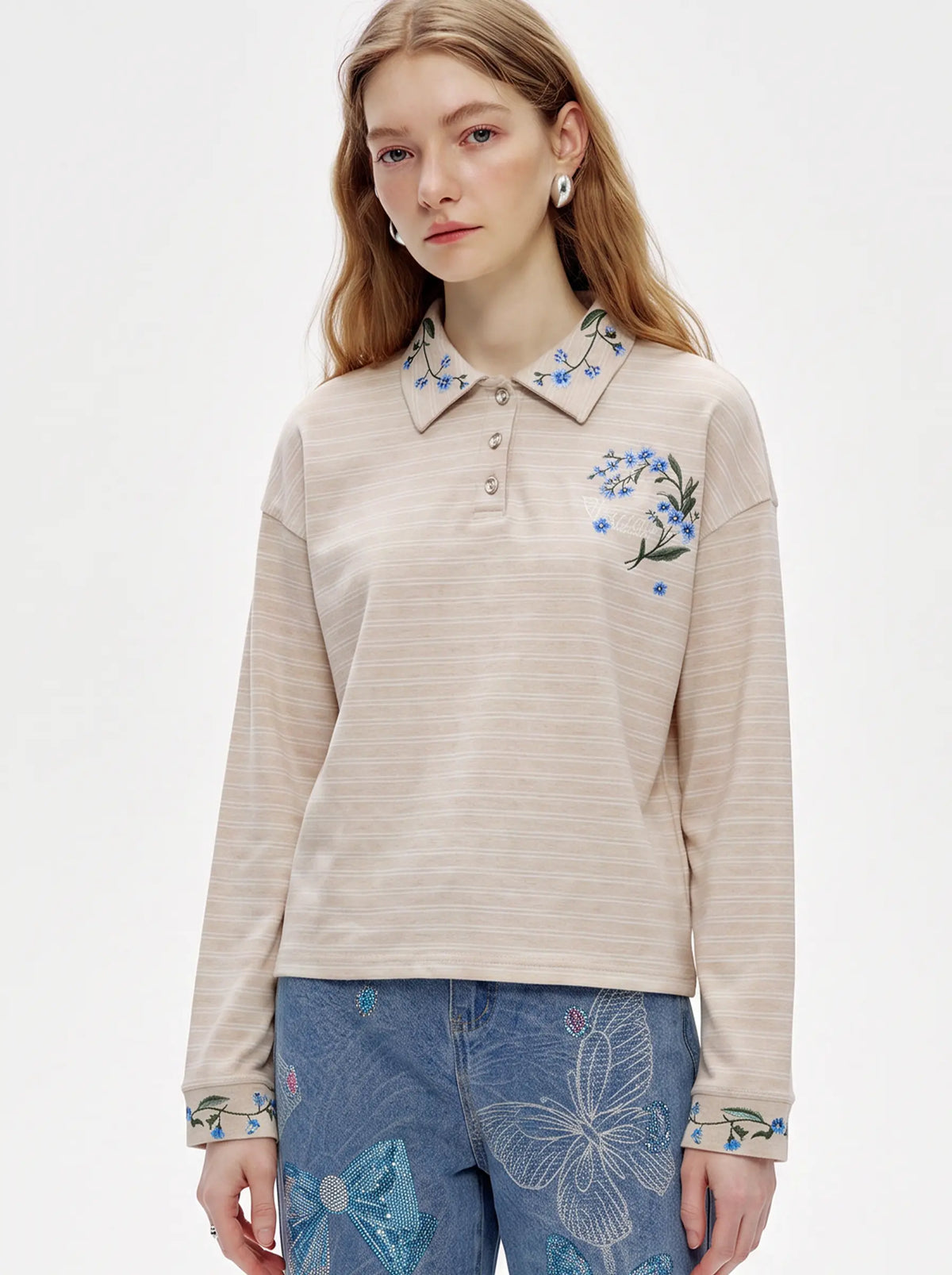 A woman wearing a beige long-sleeve polo shirt with blue floral embroidery on the collar and chest, paired with blue patterned jeans, against a plain white studio background. TCH Beige Floral Embroidered Long Sleeve Polo Shirt