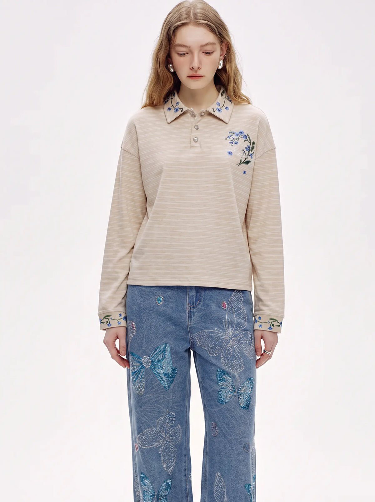 A model wearing a beige long-sleeve polo shirt with blue floral embroidery on the collar and chest, paired with blue jeans featuring a butterfly pattern, against a plain white studio background. TCH Beige Floral Embroidered Long Sleeve Polo Shirt