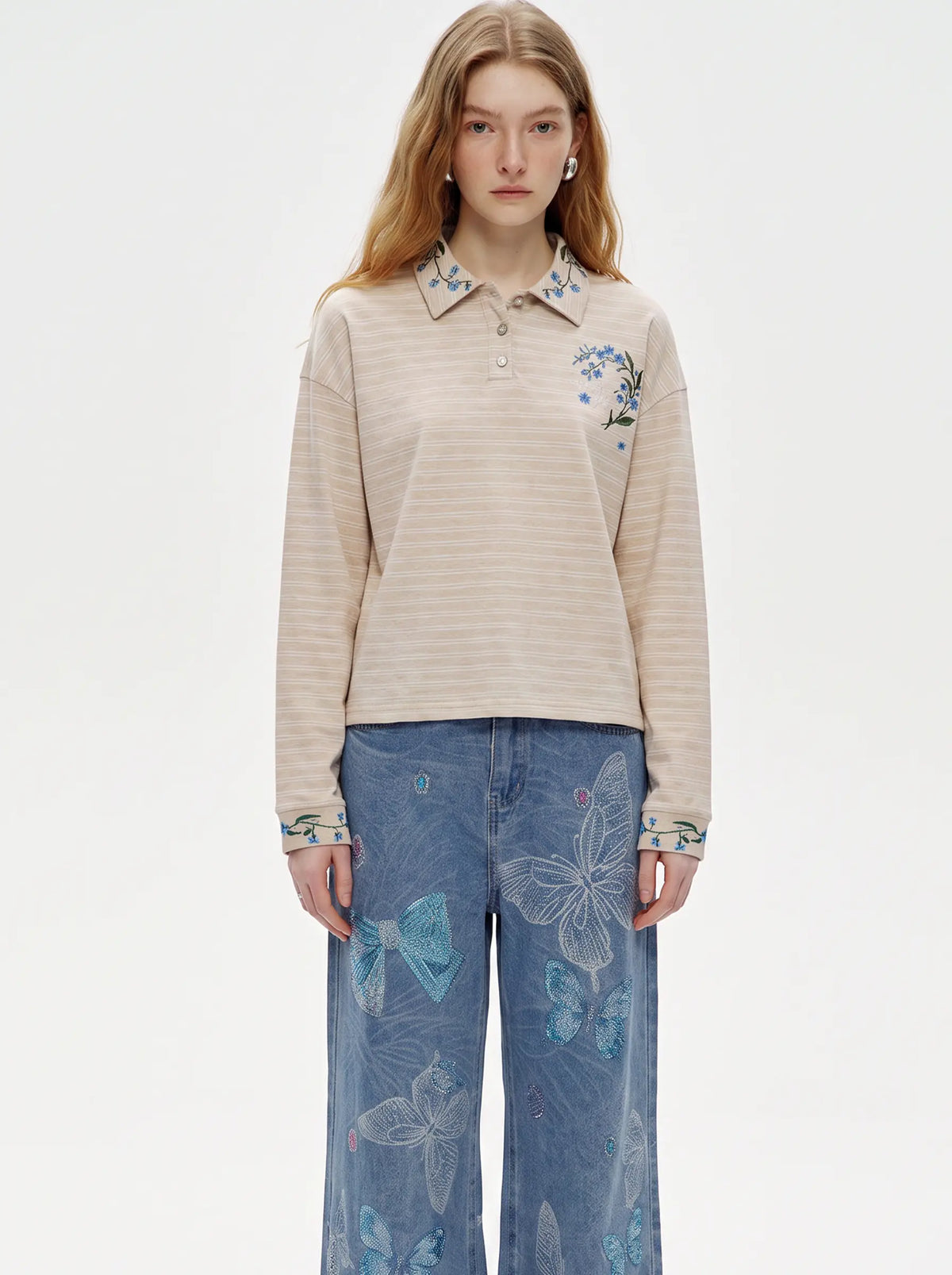A model wearing a beige and white striped long-sleeve polo shirt with blue floral embroidery on the chest and cuffs, paired with blue wide-leg jeans featuring butterfly prints, against a plain white studio background. TCH Beige Floral Embroidered Long Sleeve Polo Shirt