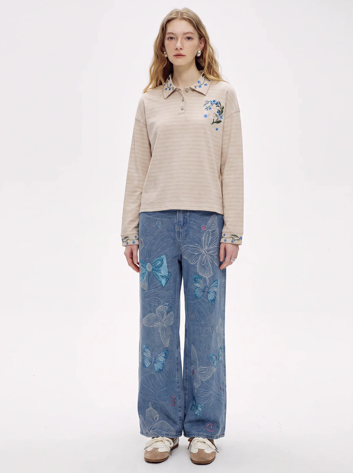 A model stands against a plain white studio background wearing a beige long-sleeve collared top with blue floral embroidery on the chest and cuffs, paired with wide-leg blue jeans featuring butterfly prints and beige shoes. TCH Beige Floral Embroidered Long Sleeve Polo Shirt