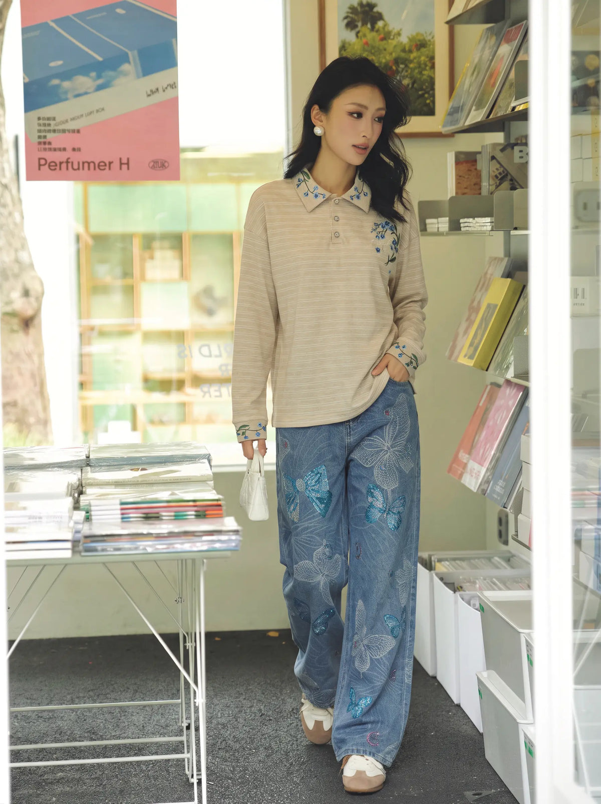A woman stands in a shop wearing a beige striped long-sleeve polo shirt with blue floral embroidery on the collar and cuffs, paired with wide-leg blue jeans featuring a large blue floral print. She is holding a small white handbag. TCH Beige Floral Embroidered Long Sleeve Polo Shirt