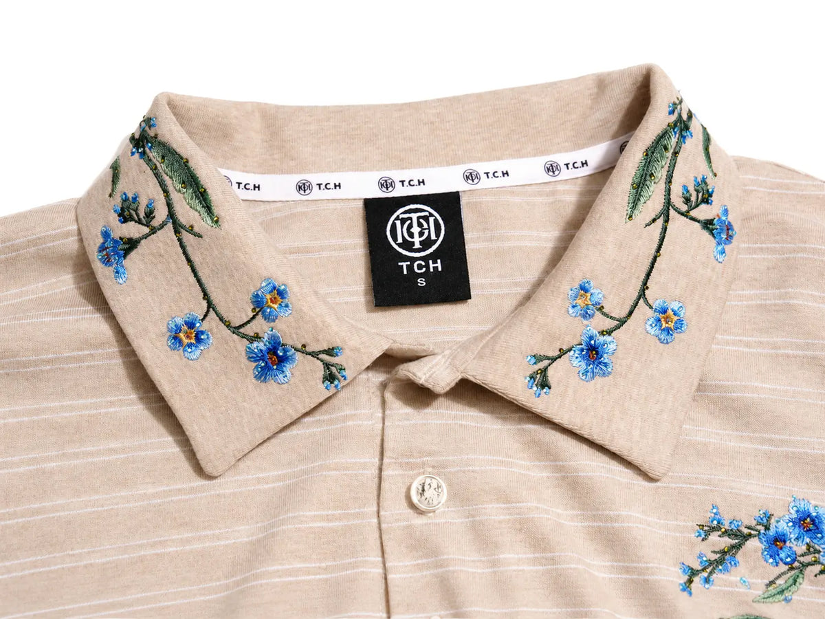 Close-up of a beige polo shirt collar with blue and green floral embroidery, featuring a black TCH brand label. TCH Beige Floral Embroidered Long Sleeve Polo Shirt