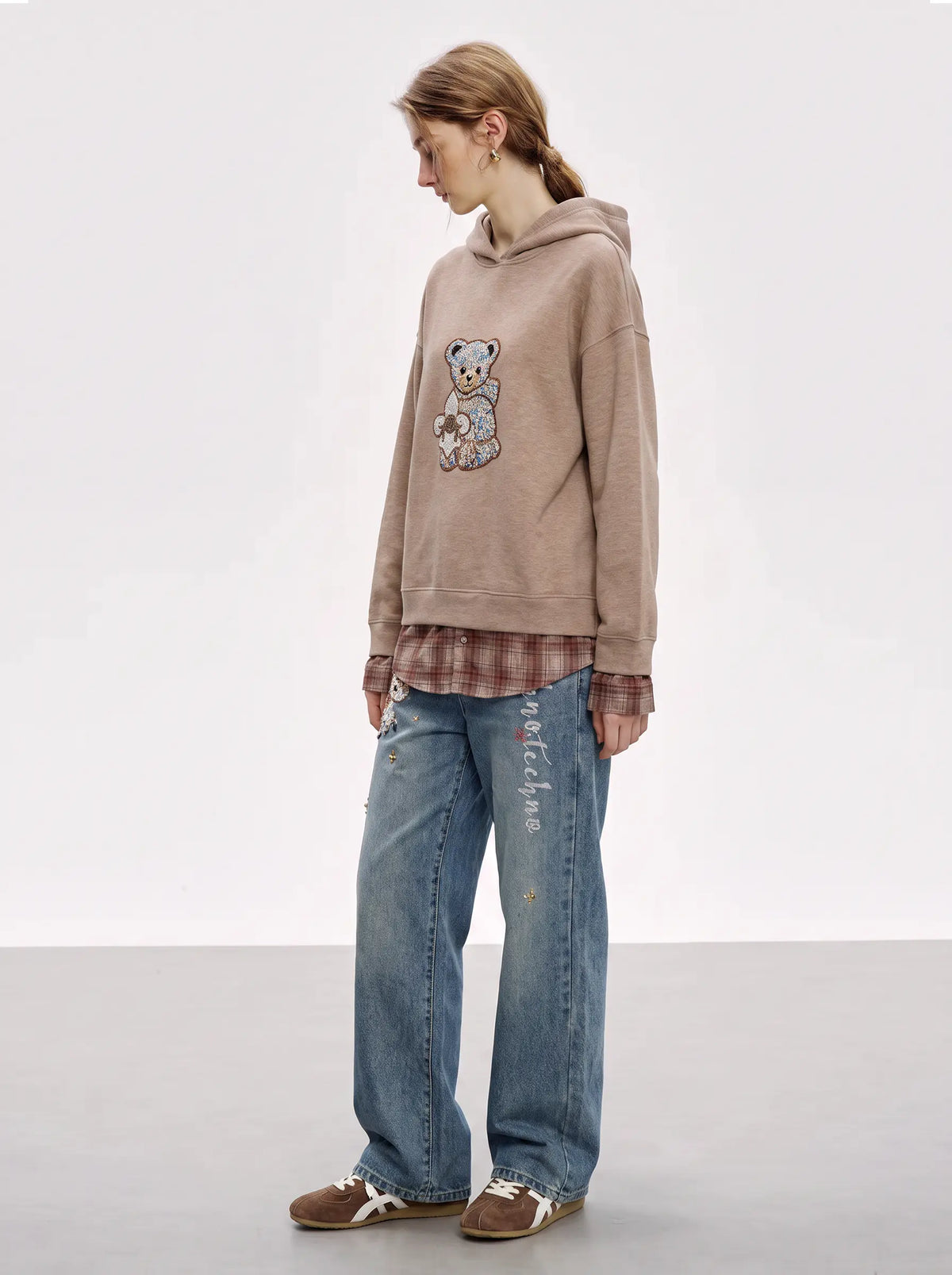 A model is shown from the side wearing a beige oversized hoodie with a teddy bear graphic, a brown plaid shirt underneath, blue jeans, and brown sneakers, set against a plain light gray studio background. TCH Beige Teddy Bear Rhinestone Layered Hoodie Sweatshirt