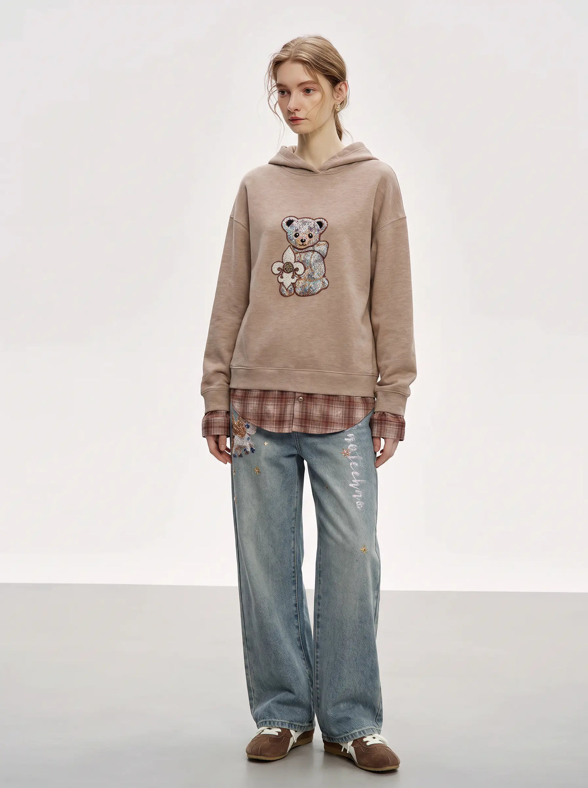 A model stands against a light gray studio background wearing a beige pullover hoodie with a teddy bear graphic, a brown plaid shirt underneath, light-wash blue jeans, and brown shoes. TCH Beige Teddy Bear Rhinestone Layered Hoodie Sweatshirt
