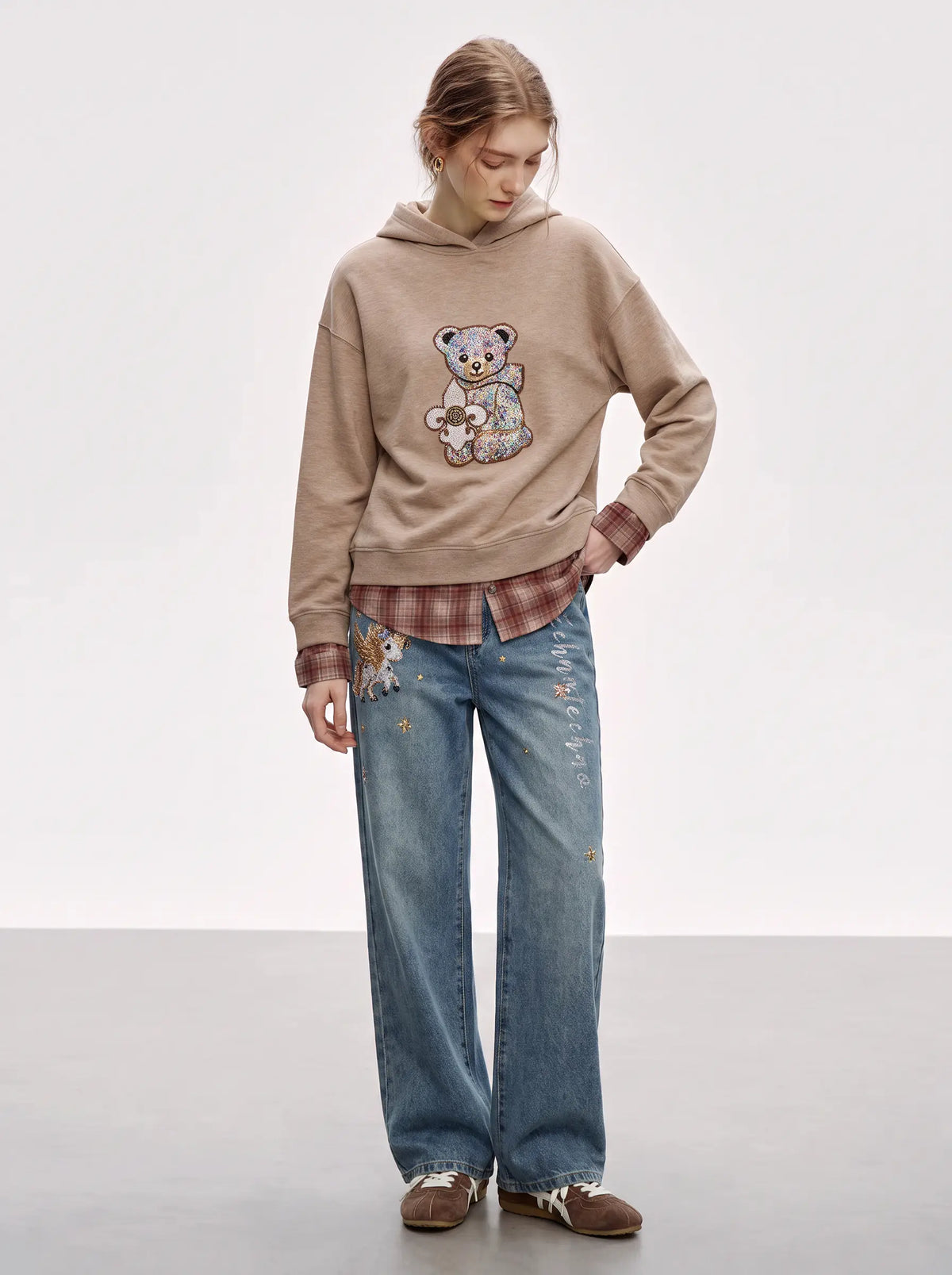 A model stands in a studio wearing a beige hoodie with a teddy bear patch, a brown plaid shirt underneath, blue jeans with embroidery, and brown shoes. TCH Beige Teddy Bear Rhinestone Layered Hoodie Sweatshirt