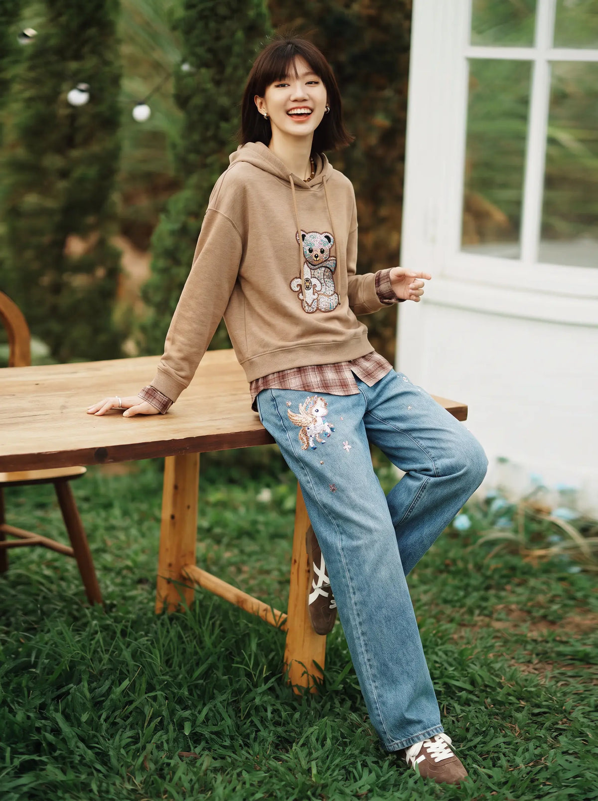 A woman smiling while leaning on a wooden table outdoors, wearing a tan hoodie with a teddy bear patch, a plaid shirt, blue jeans with floral embroidery, and brown sneakers. TCH Beige Teddy Bear Rhinestone Layered Hoodie Sweatshirt