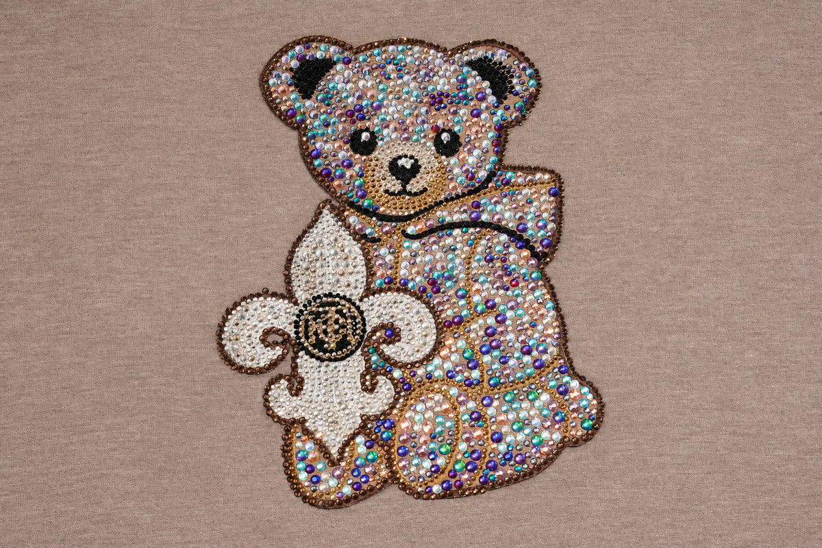 A close-up of a teddy bear and fleur-de-lis applique patch made of iridescent rhinestones on a beige fabric background. TCH Beige Teddy Bear Rhinestone Layered Hoodie Sweatshirt