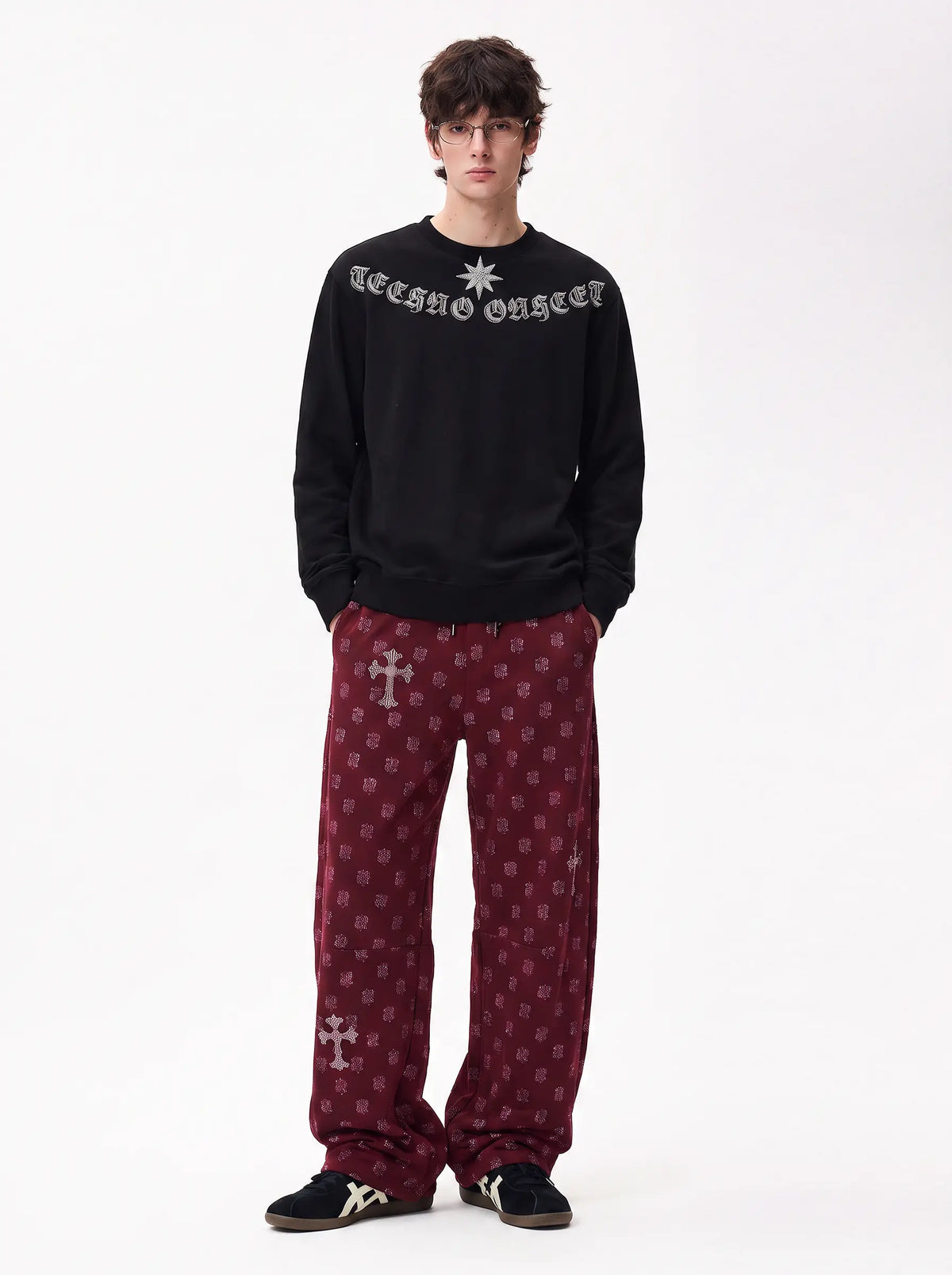A model stands against a plain white studio background wearing a black crewneck sweatshirt with 'Lethal Organism' text and a star graphic on the chest, paired with burgundy patterned pants featuring white crosses and black sneakers. TCH Black Old English Techno Graphic Sweatshirt