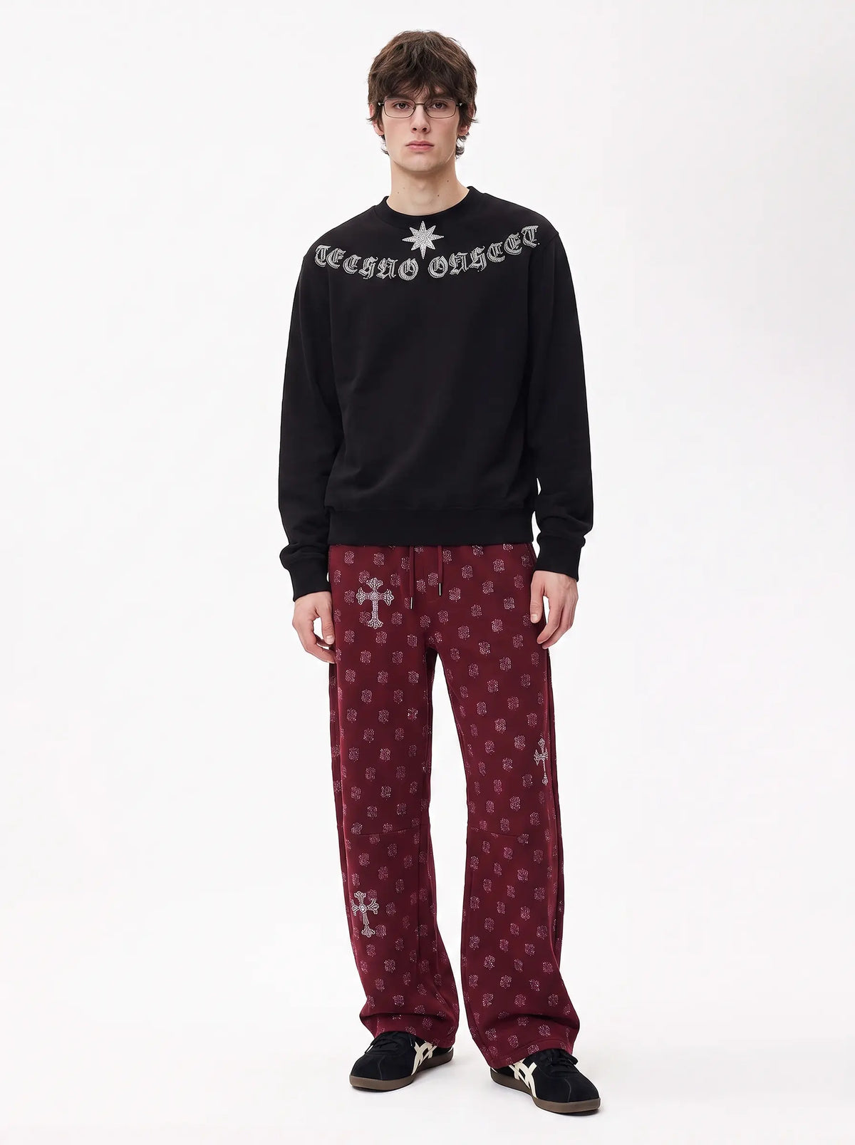 A model stands against a white studio background wearing a black crewneck sweatshirt with 'Malthusian Order' in gothic lettering and a silver star, paired with burgundy patterned pants and black sneakers. TCH Black Old English Techno Graphic Sweatshirt