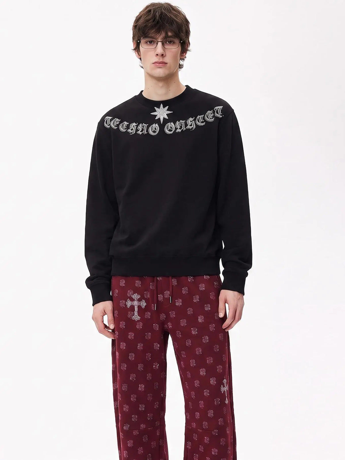 A model wearing a black crewneck sweatshirt with 'CROSS ORDER' in gothic lettering and a silver star on the chest, paired with burgundy patterned pants, against a plain white studio background. TCH Black Old English Techno Graphic Sweatshirt