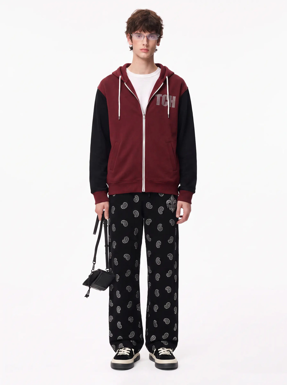 A model stands against a plain white studio background wearing a color-blocked zip-up hoodie with a burgundy body, black sleeves, white drawstrings, and a white graphic print on the left chest. The hoodie is layered over a white t-shirt and paired with black paisley-print pants, black sneakers, and a small black handbag. TCH Black Paisley Print Wide-Leg Sweatpants
