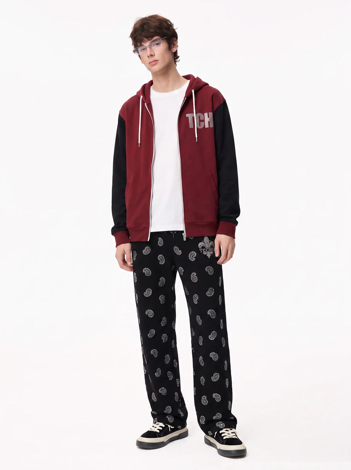 A model stands against a plain white studio background wearing a maroon and black zip-up hoodie with 'TCH' printed on the chest, a white t-shirt, black patterned sweatpants, and black sneakers. TCH Black Paisley Print Wide-Leg Sweatpants