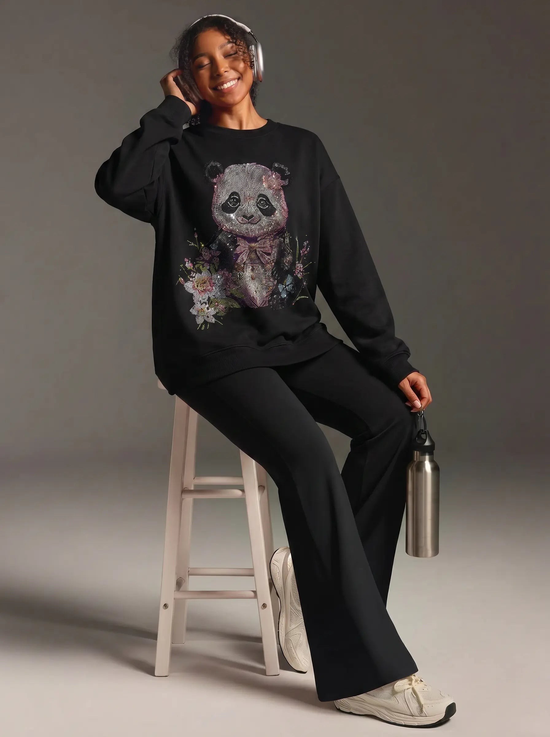 A woman wearing a black crewneck sweatshirt with a large floral panda graphic and matching black pants, sitting on a white stool and holding a silver water bottle against a gray studio background. TCH Black Panda Rhinestone Sweatshirt