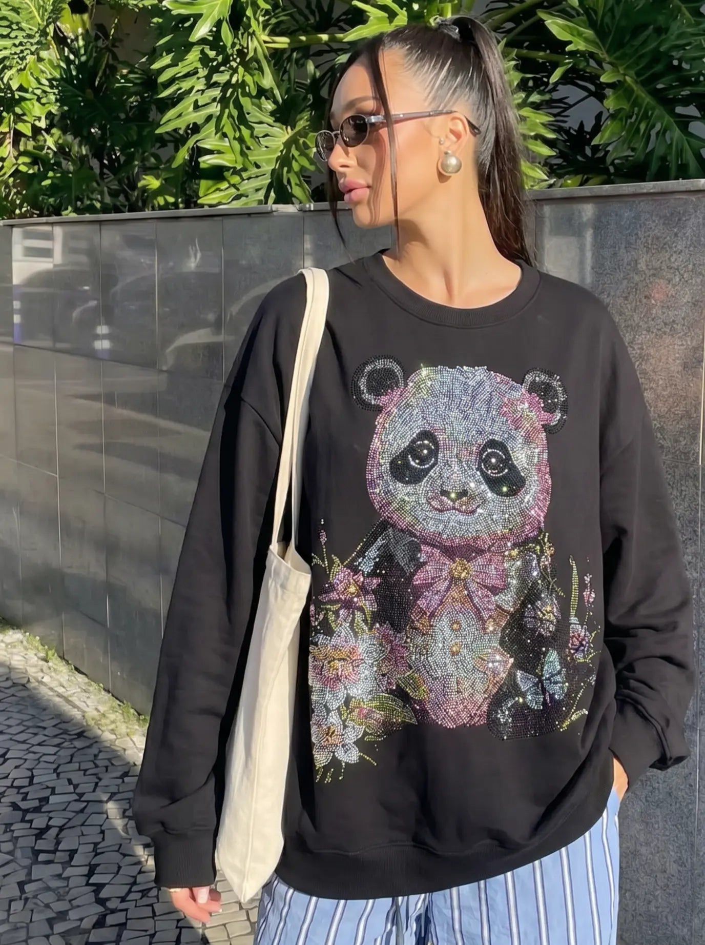 A woman wearing a black crewneck sweatshirt with a large, glittery panda graphic on the front, paired with blue and white striped pants and a beige canvas tote bag, standing outdoors against a dark wall and green foliage. TCH Black Panda Rhinestone Sweatshirt