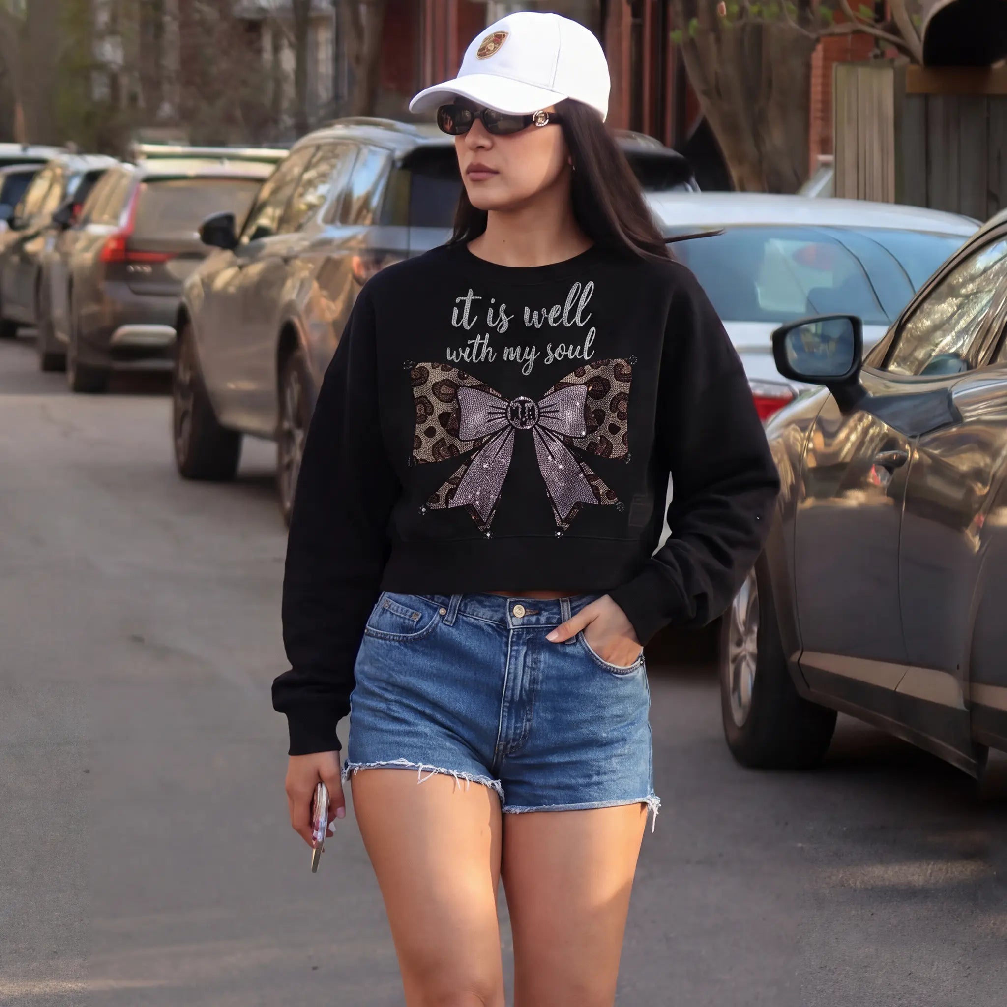 Woman wearing a black sweatshirt with a bow design and text, denim shorts, and a white cap in an urban setting. TCH Black Rhinestone Bow Cropped Sweatshirt