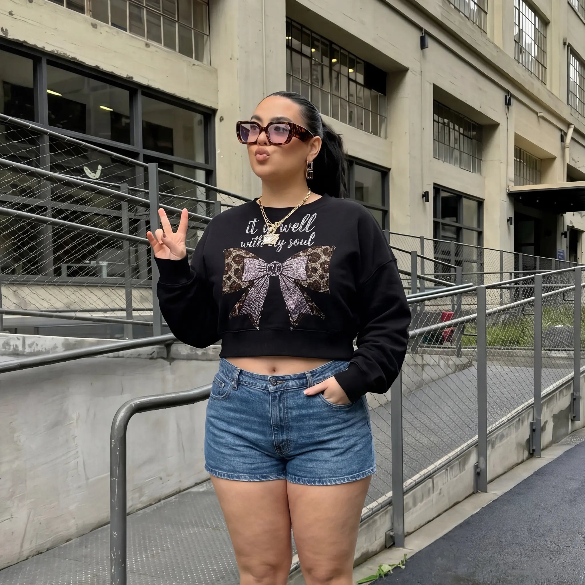 Woman wearing a black sweater with a leopard print bow design and denim shorts, standing in an urban setting. TCH Black Rhinestone Bow Cropped Sweatshirt