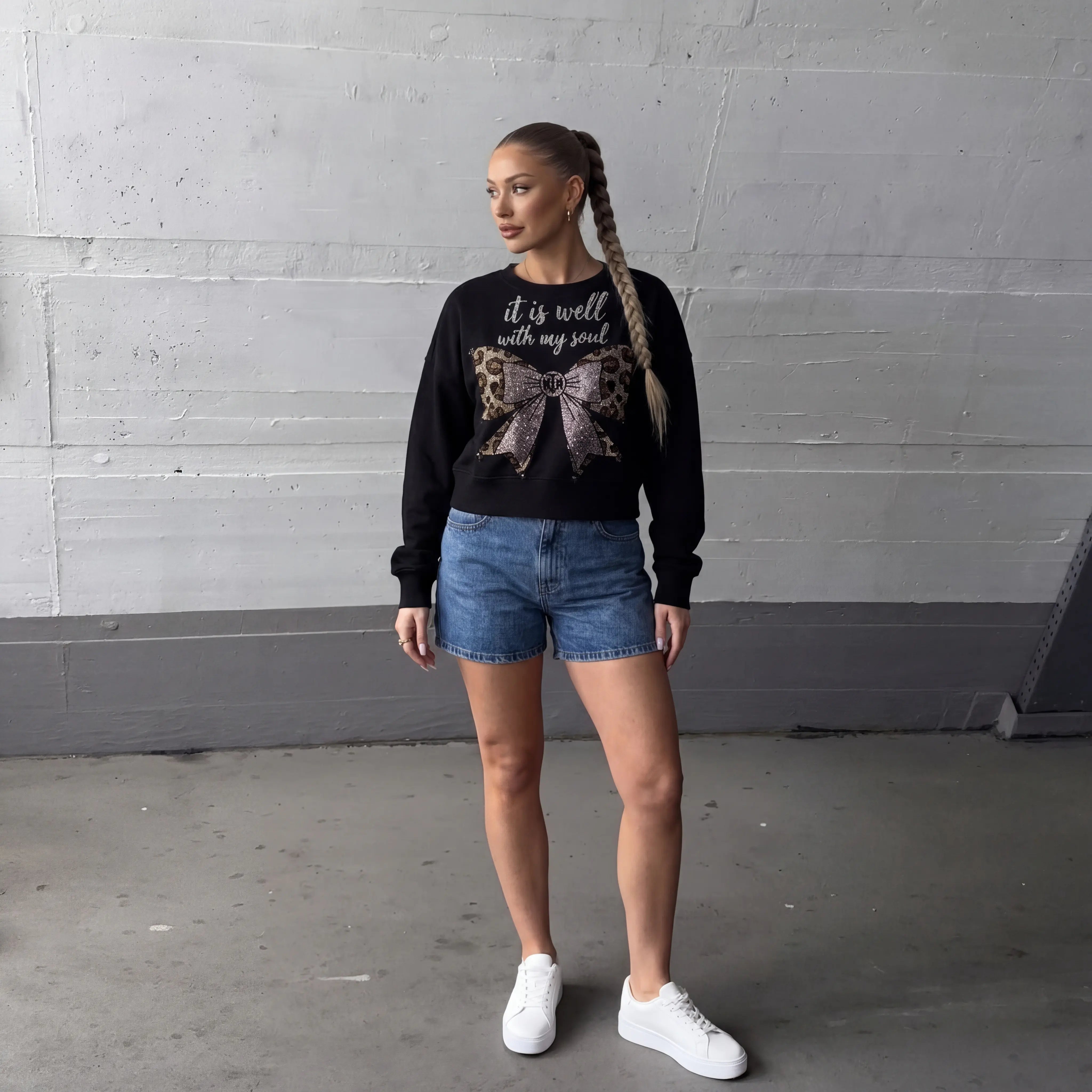 Woman wearing a black sweatshirt with text and a bow design, denim shorts, and white sneakers against a gray wall. TCH Black Rhinestone Bow Cropped Sweatshirt
