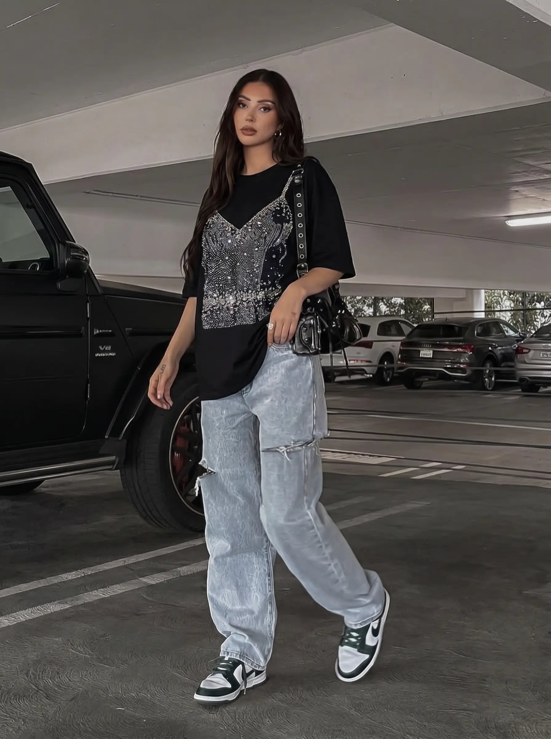 A woman walking in a parking garage wearing a black t-shirt with a silver sequined vest, light-wash distressed jeans, and black and white sneakers, carrying a black shoulder bag. TCH Black Rhinestone Corset Graphic T-Shirt