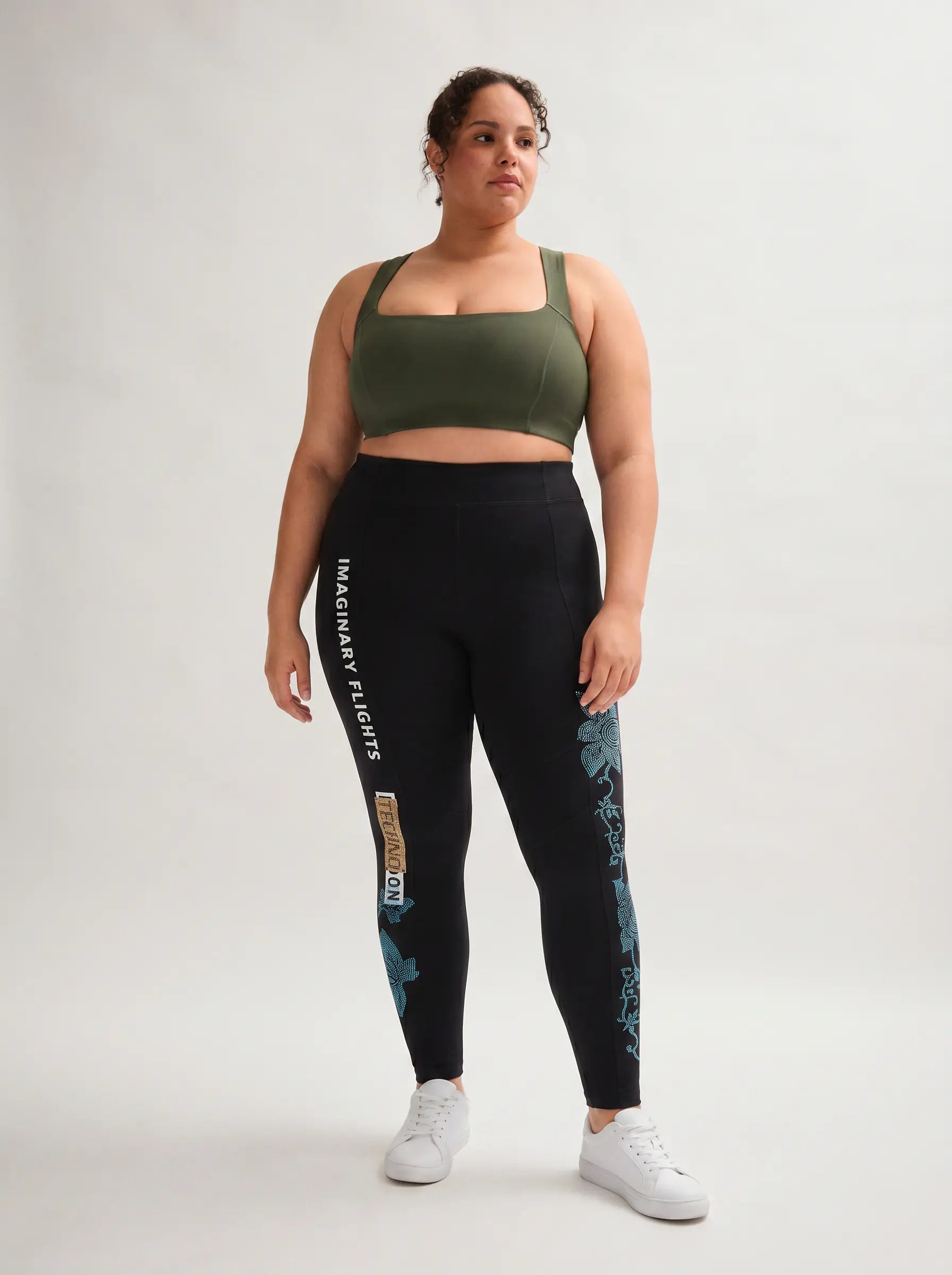 A woman modeling a matching activewear set consisting of an olive green sports bra and black leggings with teal floral patterns and white text, paired with white sneakers, against a light gray studio background. TCH Black Rhinestone Floral Leggings