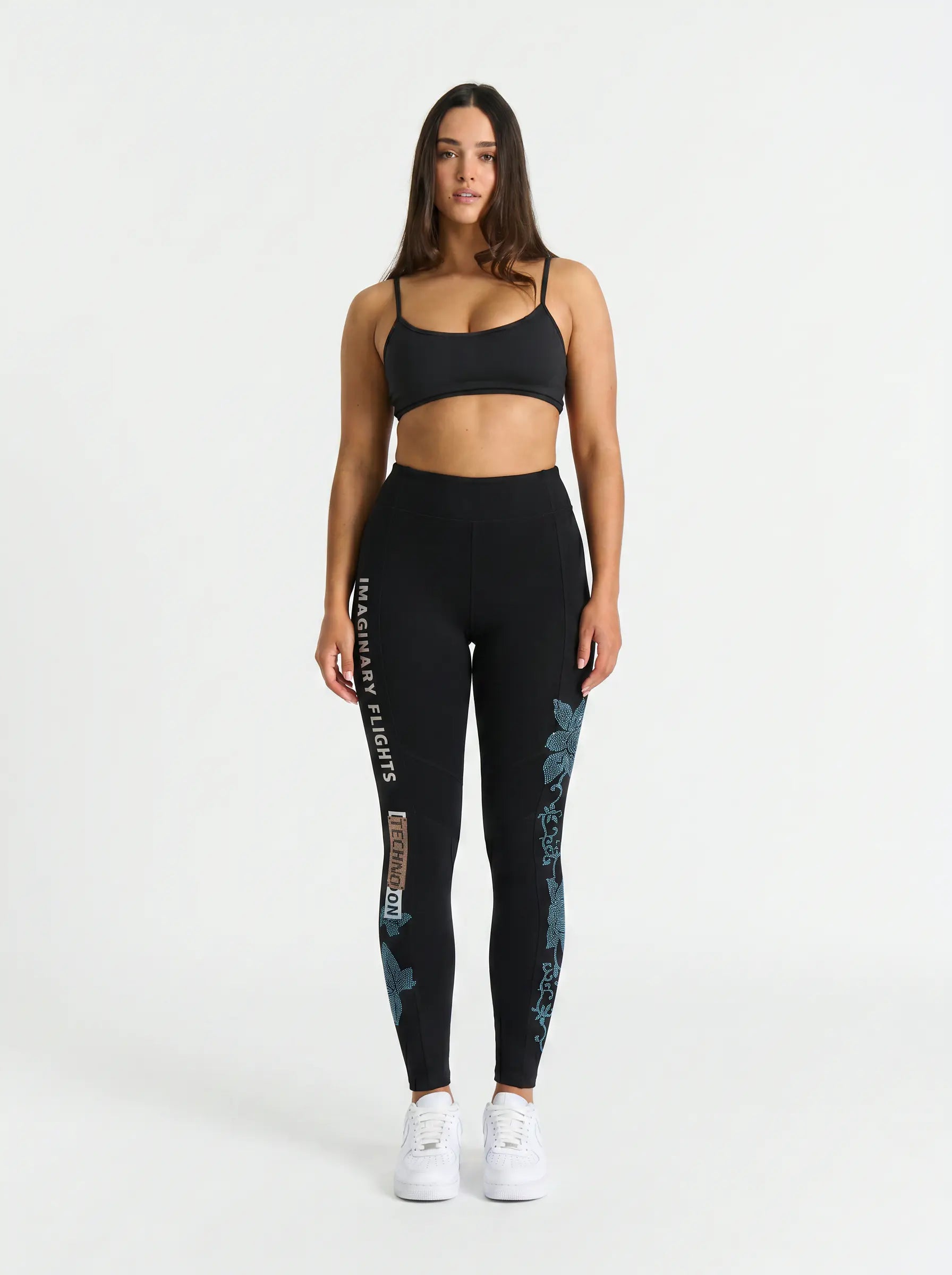 A woman modeling a black activewear set consisting of a sports bra and high-waisted leggings with teal floral patterns and white text, paired with white sneakers, against a plain white studio background. TCH Black Rhinestone Floral Leggings