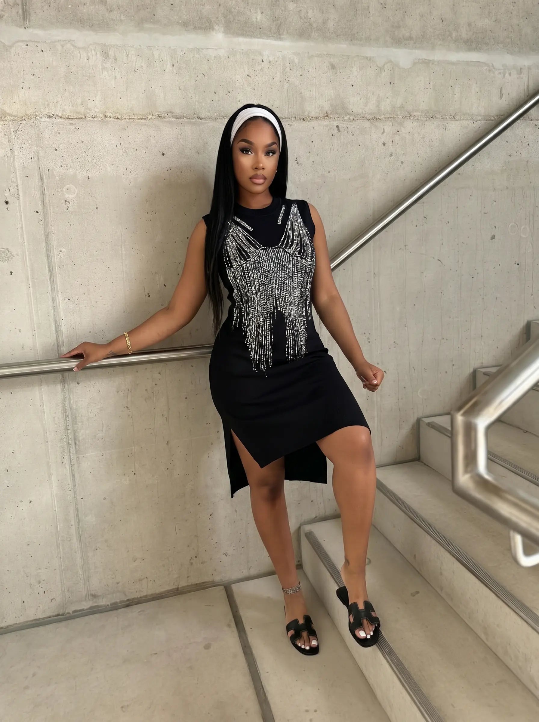 A woman posing on concrete stairs wearing a black sleeveless dress with a silver beaded fringe chest detail, a black hijab, and black flat sandals. TCH Black Rhinestone Fringe Detail Long Sleeve Mini Dress