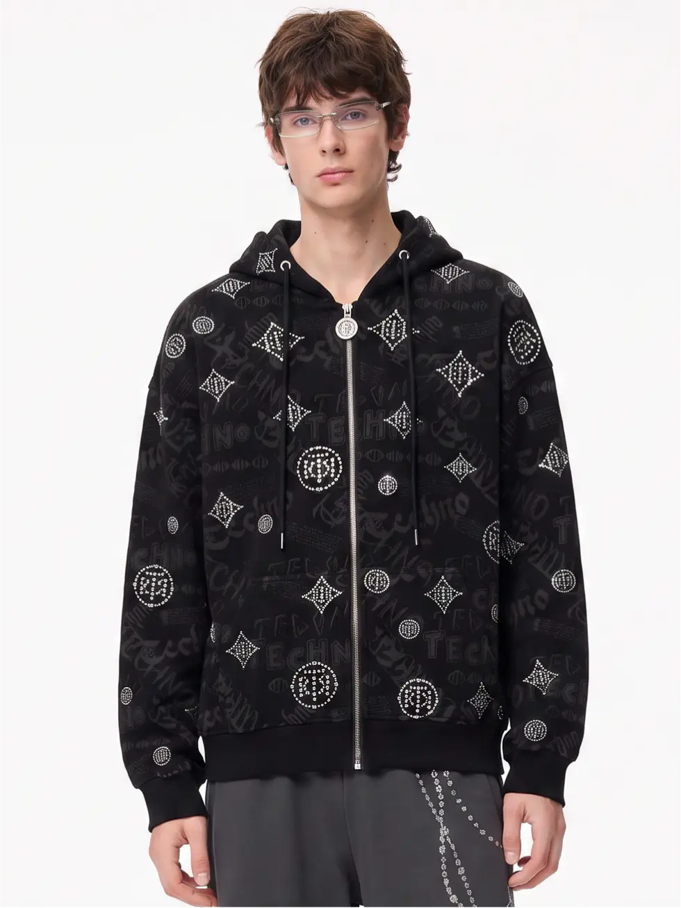 A young man wearing a black zip-up hoodie with an all-over white graffiti-style monogram pattern, paired with gray sweatpants, against a plain white studio background. TCH Black Rhinestone Monogram Zip Up Hoodie Jacket