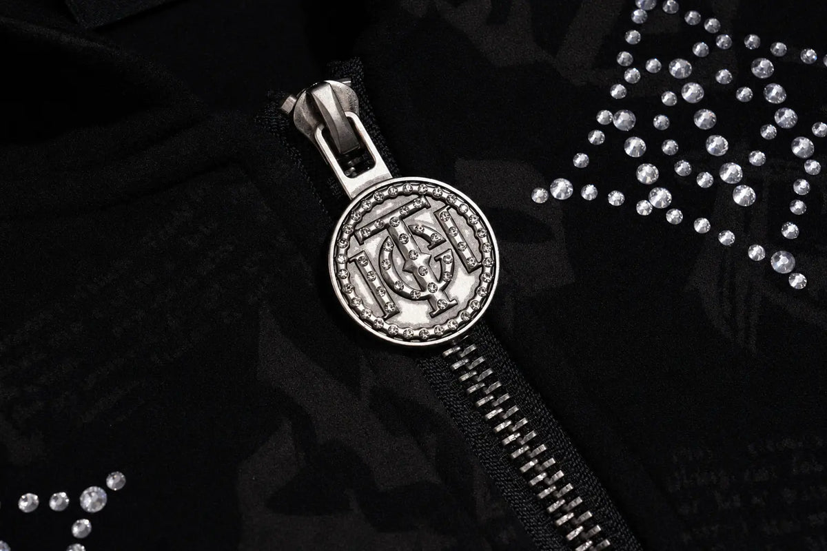 A close-up of a black zippered garment featuring a prominent round silver-tone metal zipper pull with an embossed monogram design, accented with rhinestone embellishments on the black fabric. TCH Black Rhinestone Monogram Zip Up Hoodie Jacket