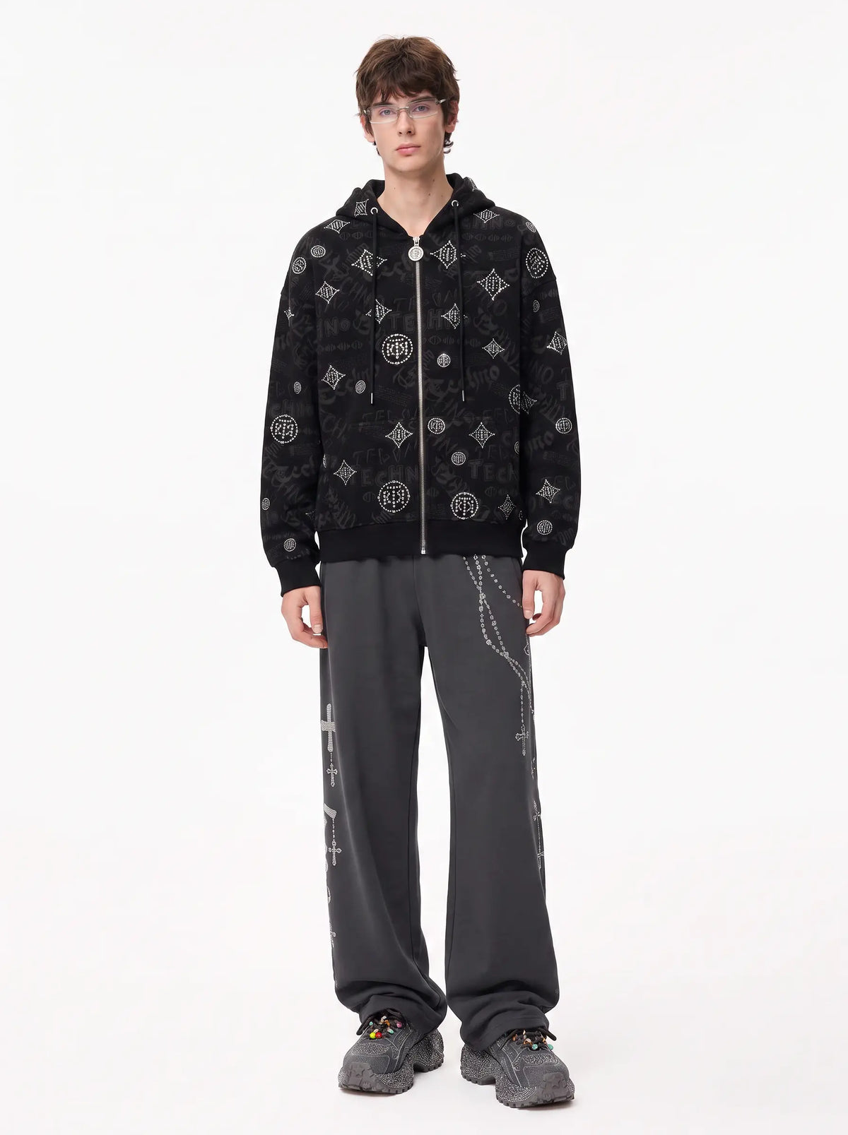 A model stands against a plain white studio background wearing a black zip-up hoodie with a white monogram-style pattern, dark gray sweatpants, and gray sneakers. TCH Black Rhinestone Monogram Zip Up Hoodie Jacket