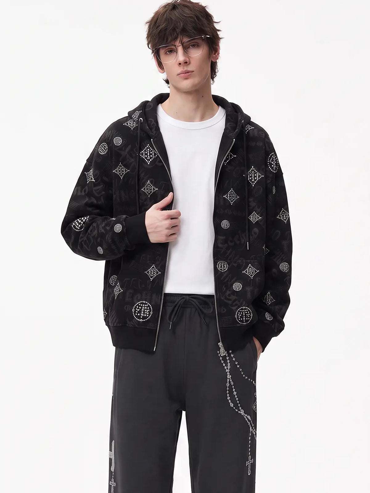 A model wearing a black zip-up hoodie with an all-over silver graphic pattern, styled over a white t-shirt and dark sweatpants against a plain white studio background. TCH Black Rhinestone Monogram Zip Up Hoodie Jacket