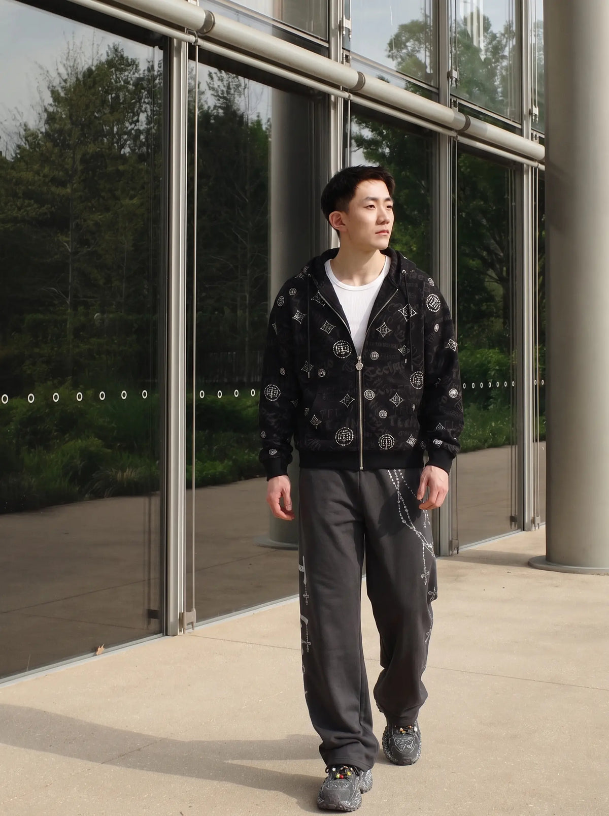 A man wearing a black zip-up hoodie with a white geometric pattern, dark gray cargo pants, and gray sneakers, standing in front of a modern glass building. TCH Black Rhinestone Monogram Zip Up Hoodie Jacket