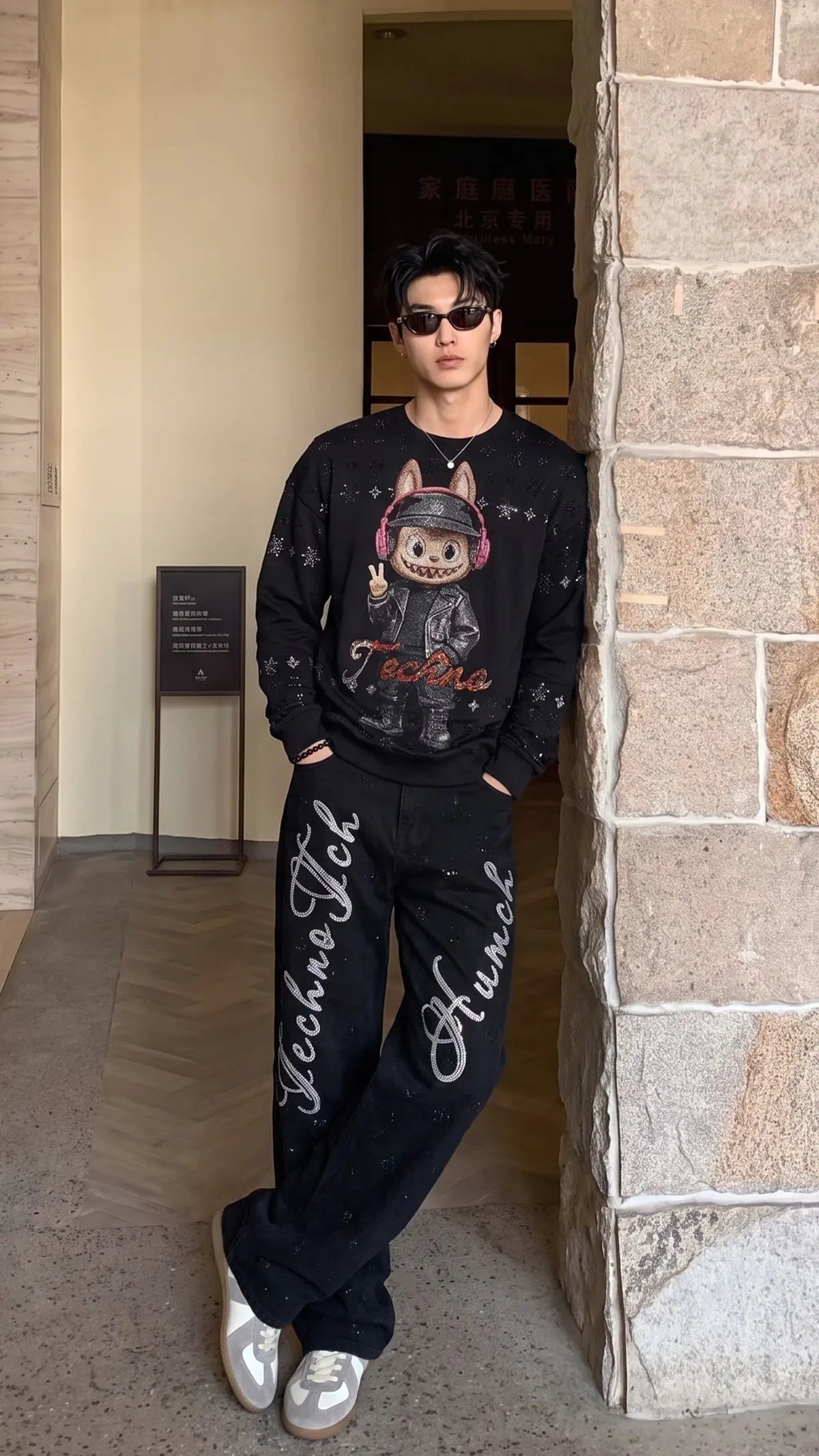 A man wearing a black graphic sweatshirt with a cartoon dog design and matching black pants with white script text, standing in a stone-walled indoor corridor. TCH Black Techno Bunny Rhinestone Graphic Sweatshirt
