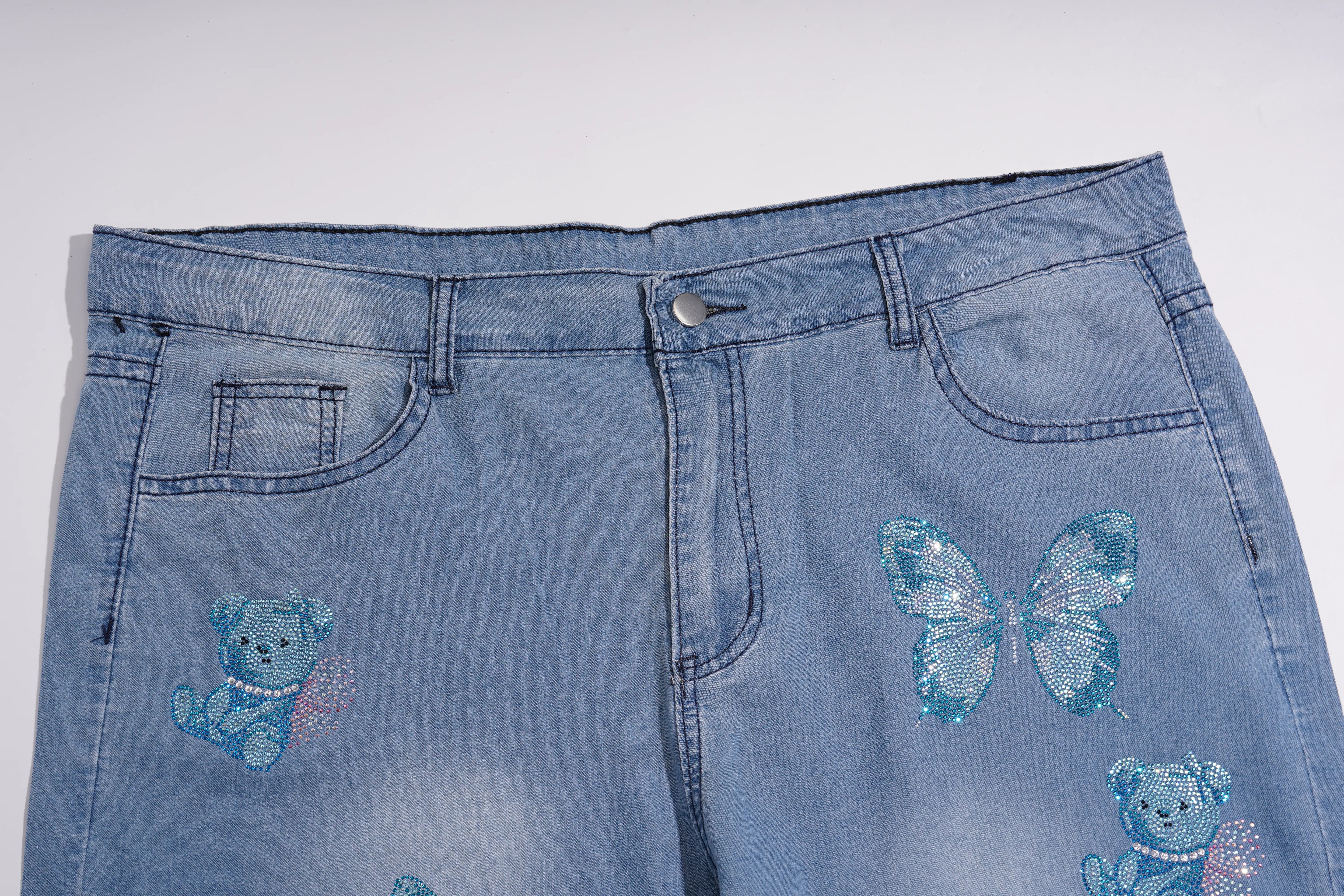 Light blue denim jeans featuring sequined blue teddy bear and butterfly designs on the front, photographed against a plain light gray background. TCH Blue Butterfly & Teddy Bear Lace Hem Denim Jeans