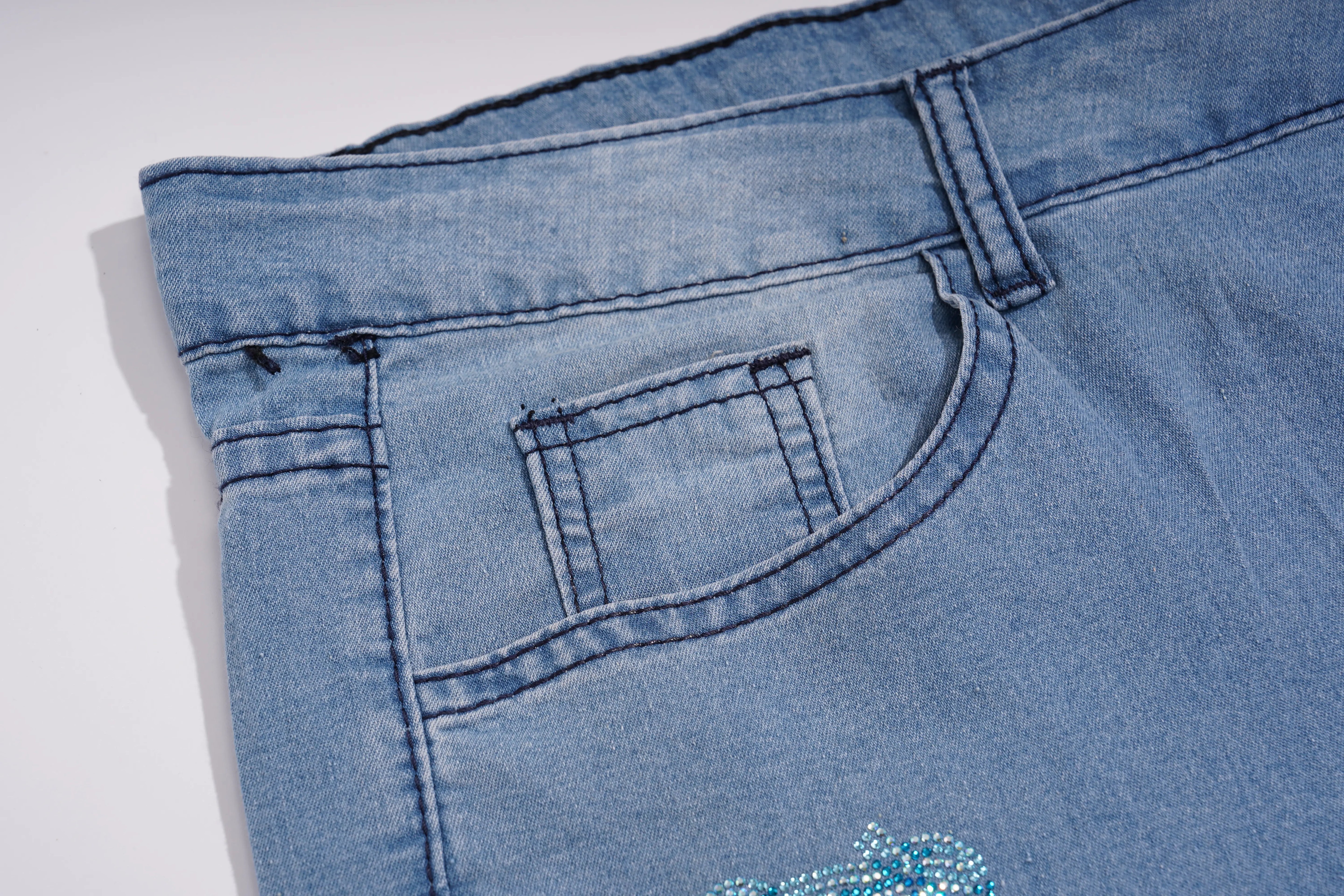 Close-up of a pair of light blue denim jeans showing the back pocket, waistband, and a small embellished detail near the lower leg. TCH Blue Butterfly & Teddy Bear Lace Hem Denim Jeans