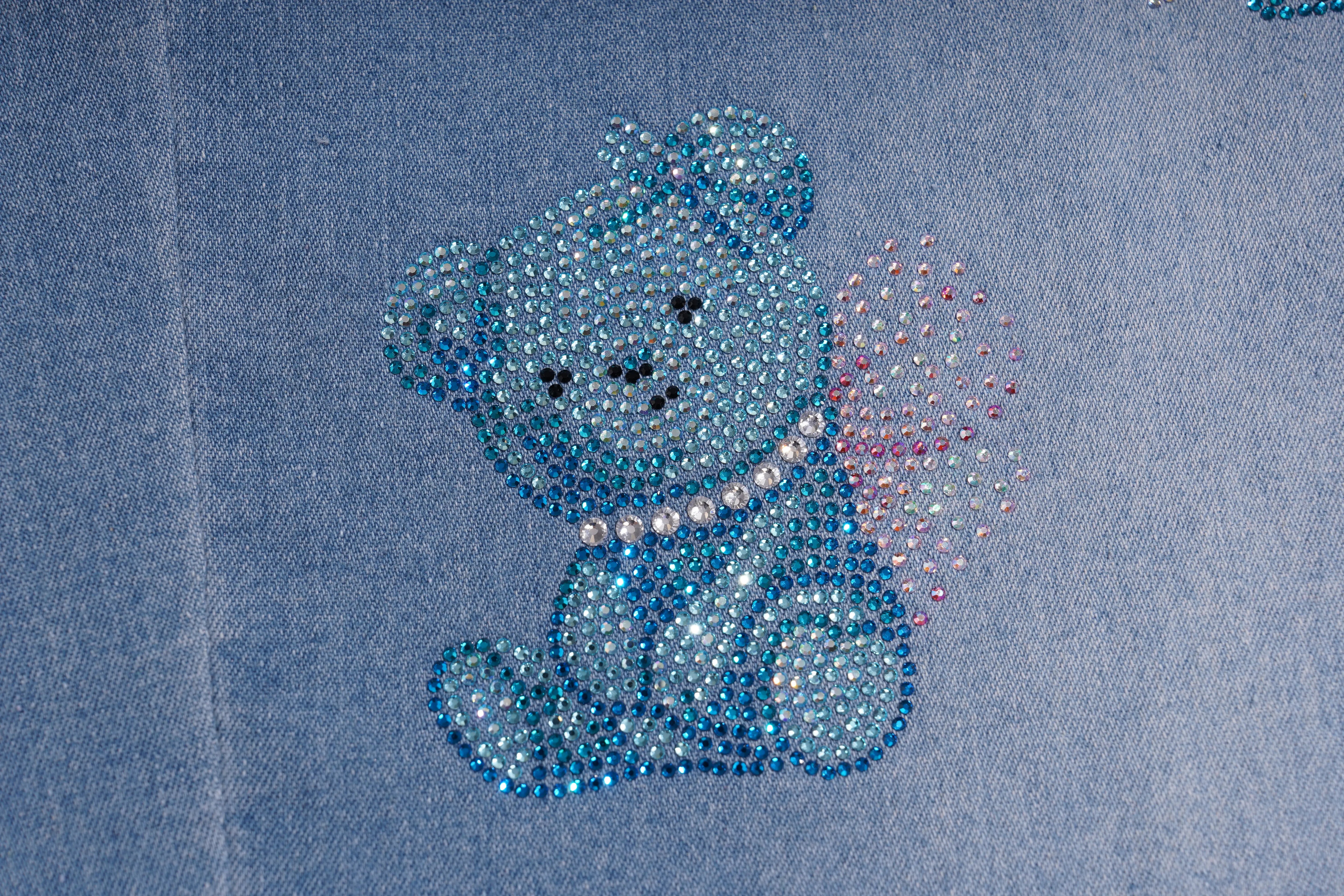 A close-up of a blue denim garment featuring an embroidered or applique teddy bear design made with rhinestones. The bear is light blue with a white pearl-like collar and a pink heart-shaped tail, set against the blue fabric background. TCH Blue Butterfly & Teddy Bear Lace Hem Denim Jeans