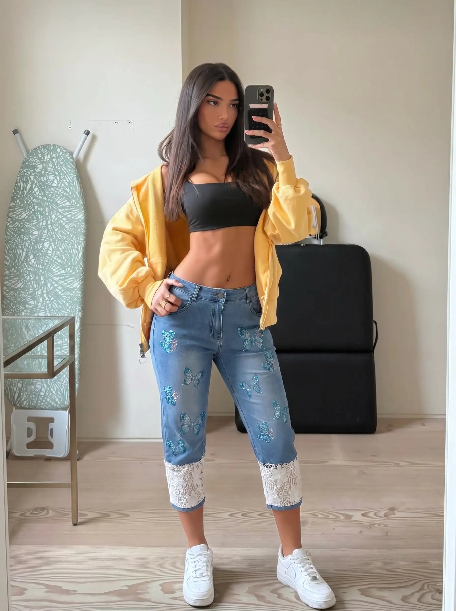 A woman takes a mirror selfie in a room, wearing a yellow open-front jacket, a black crop top, distressed blue jeans with light blue floral embroidery and white lace cuffs, and white sneakers. TCH Blue Butterfly & Teddy Bear Lace Hem Denim Jeans