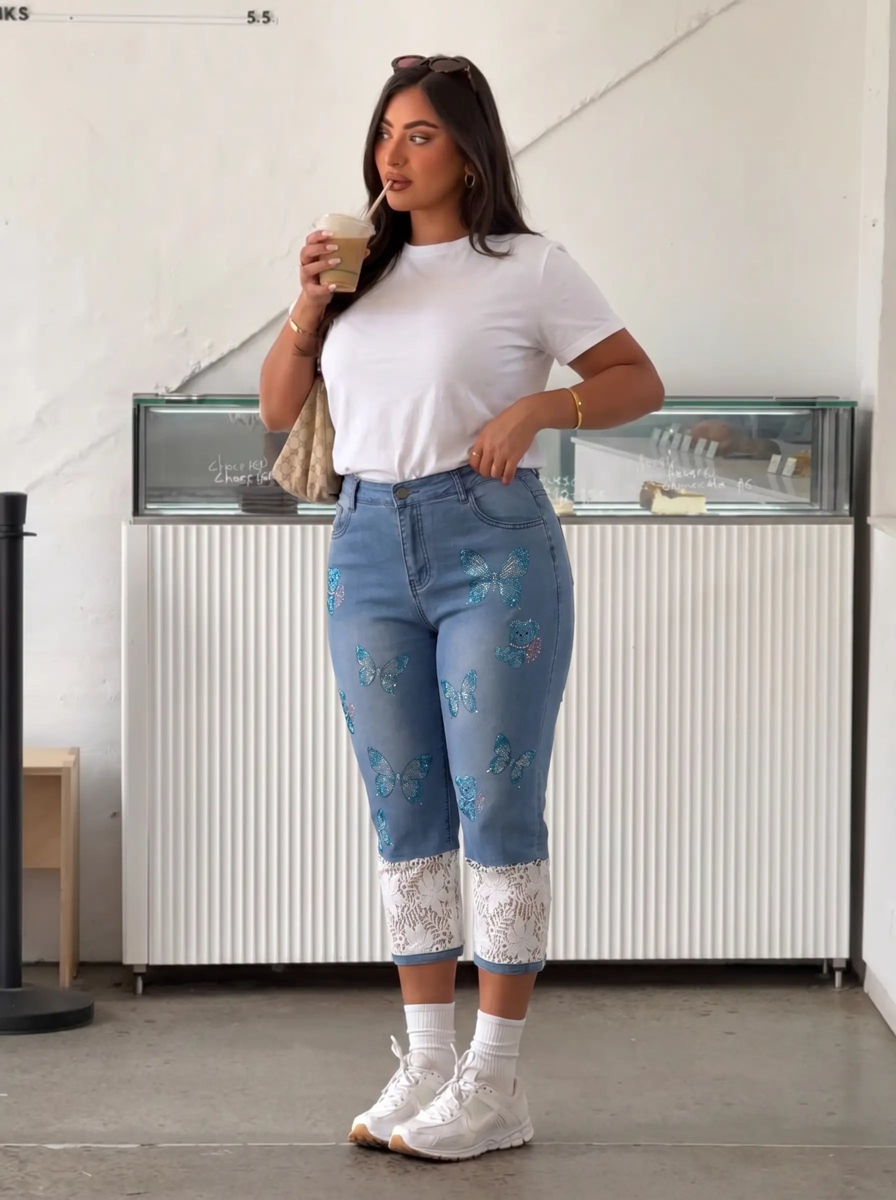 A woman modeling a pair of light blue denim jeans with blue floral embroidery and white lace cuffs, styled with a white t-shirt and white sneakers in a cafe setting. TCH Blue Butterfly & Teddy Bear Lace Hem Denim Jeans