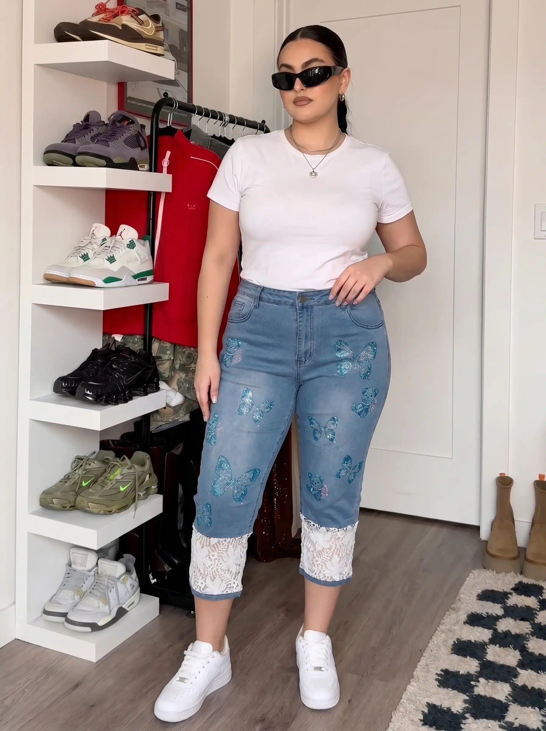 A woman modeling a pair of light blue cropped denim jeans with blue butterfly prints and white lace hem detailing, styled with a white t-shirt and white sneakers in a room with a shoe rack. TCH Blue Butterfly & Teddy Bear Lace Hem Denim Jeans