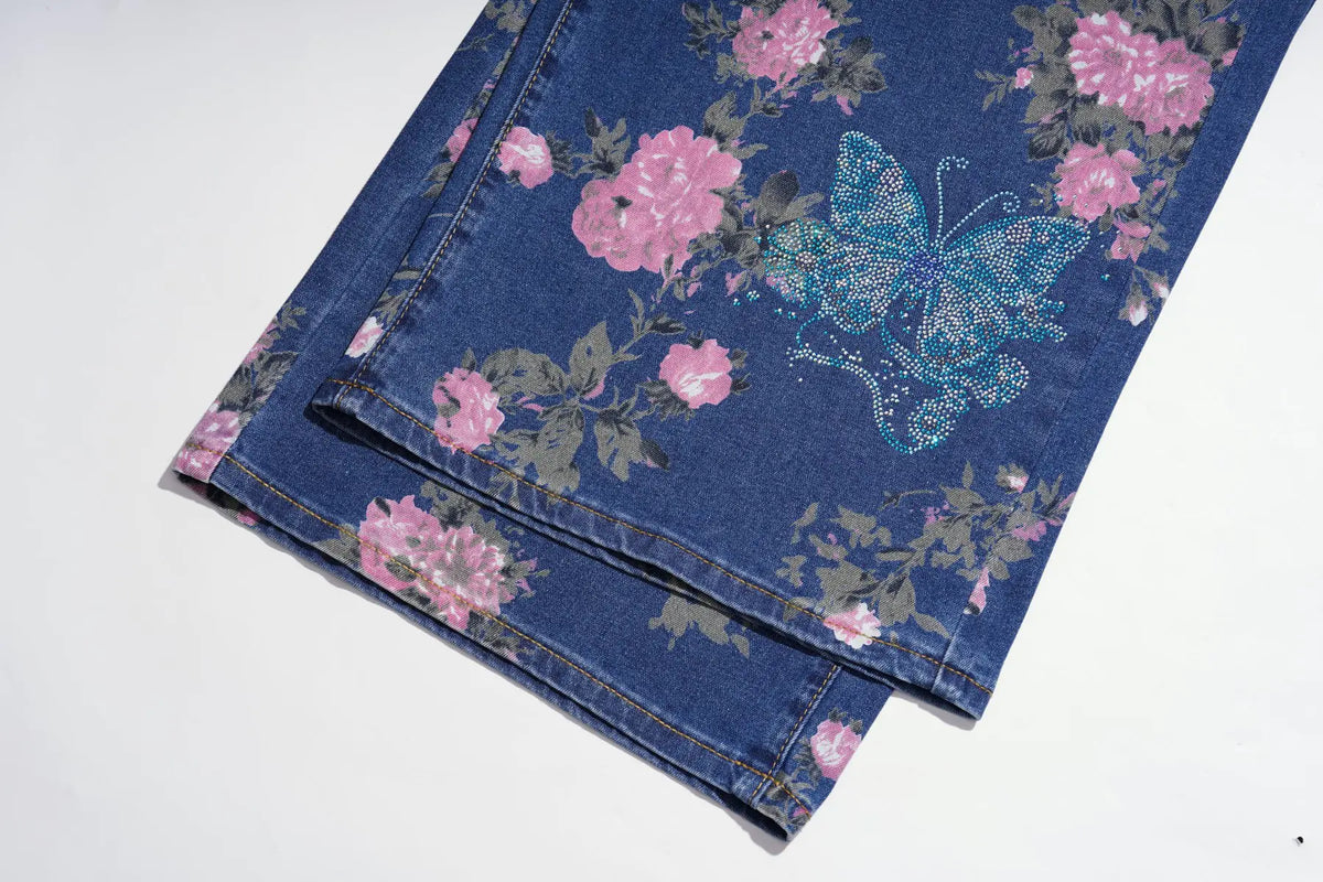 A close-up of folded blue denim jeans featuring a pink floral print and a glittery blue butterfly design on a white background. TCH Blue Floral Butterfly Rhinestone Wide-Leg Jeans