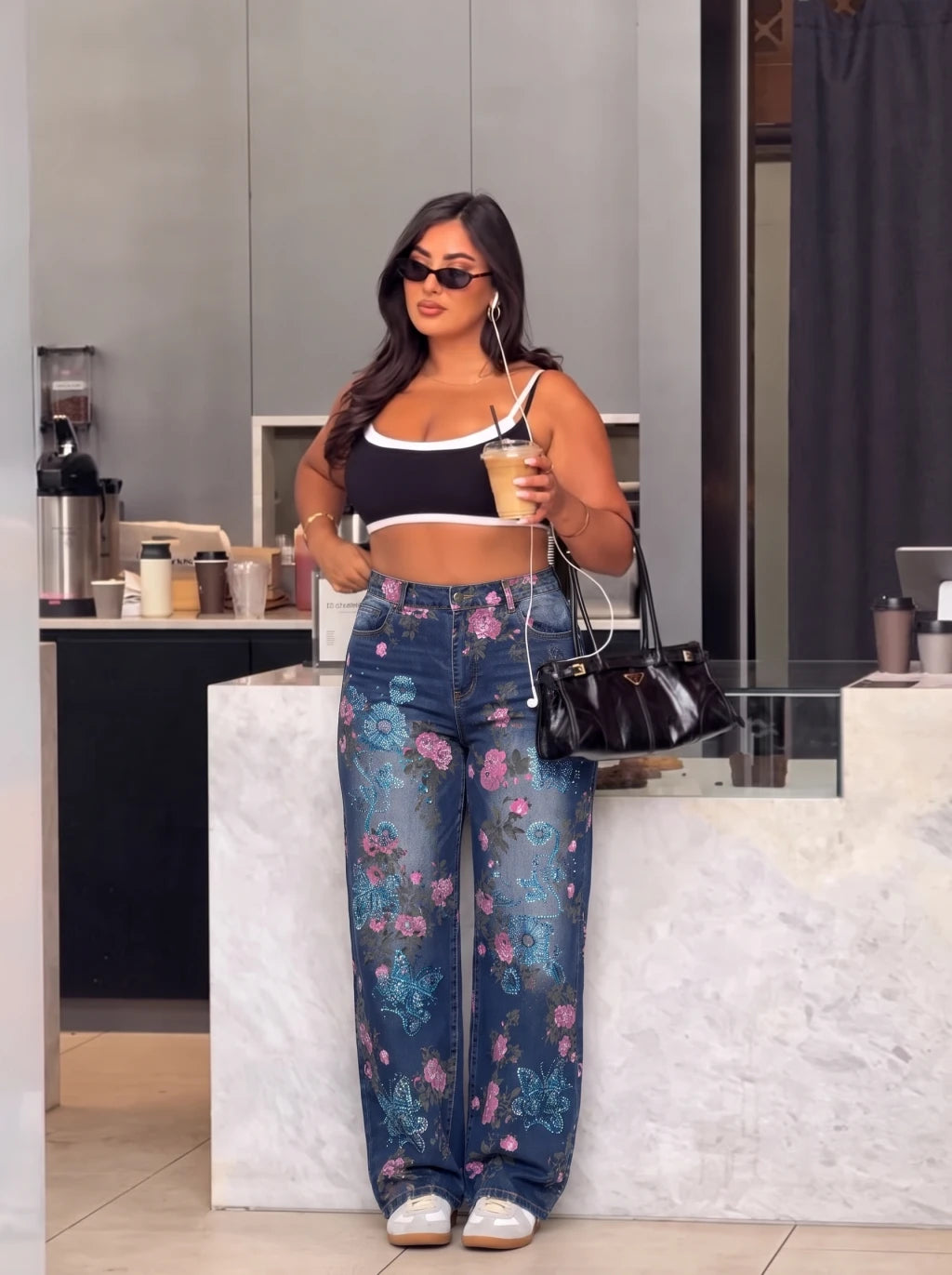 A woman stands in a modern cafe setting wearing a black and white crop top, blue floral wide-leg jeans, white sneakers, and carrying a black handbag. TCH Blue Floral Butterfly Rhinestone Wide-Leg Jeans