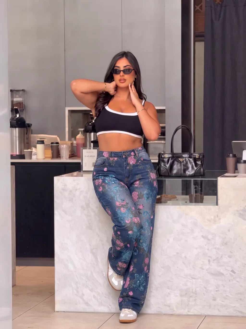 A woman posing in a modern kitchen setting, wearing a black crop top with white trim and high-waisted, wide-leg blue jeans featuring a pink and purple floral pattern. TCH Blue Floral Butterfly Rhinestone Wide-Leg Jeans